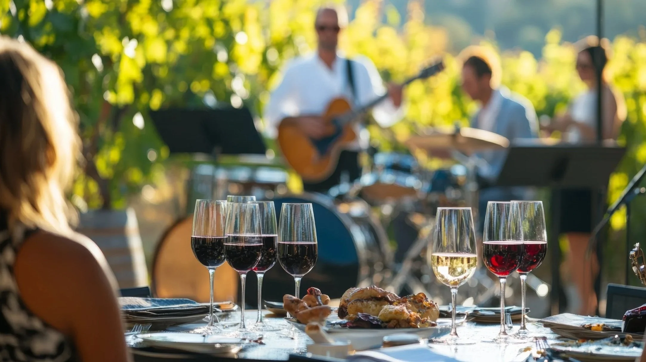 Multiple glasses of red and white wine on a table outdoors, with a blurred background of musicians playing guitars and crowds at a gathering or event.