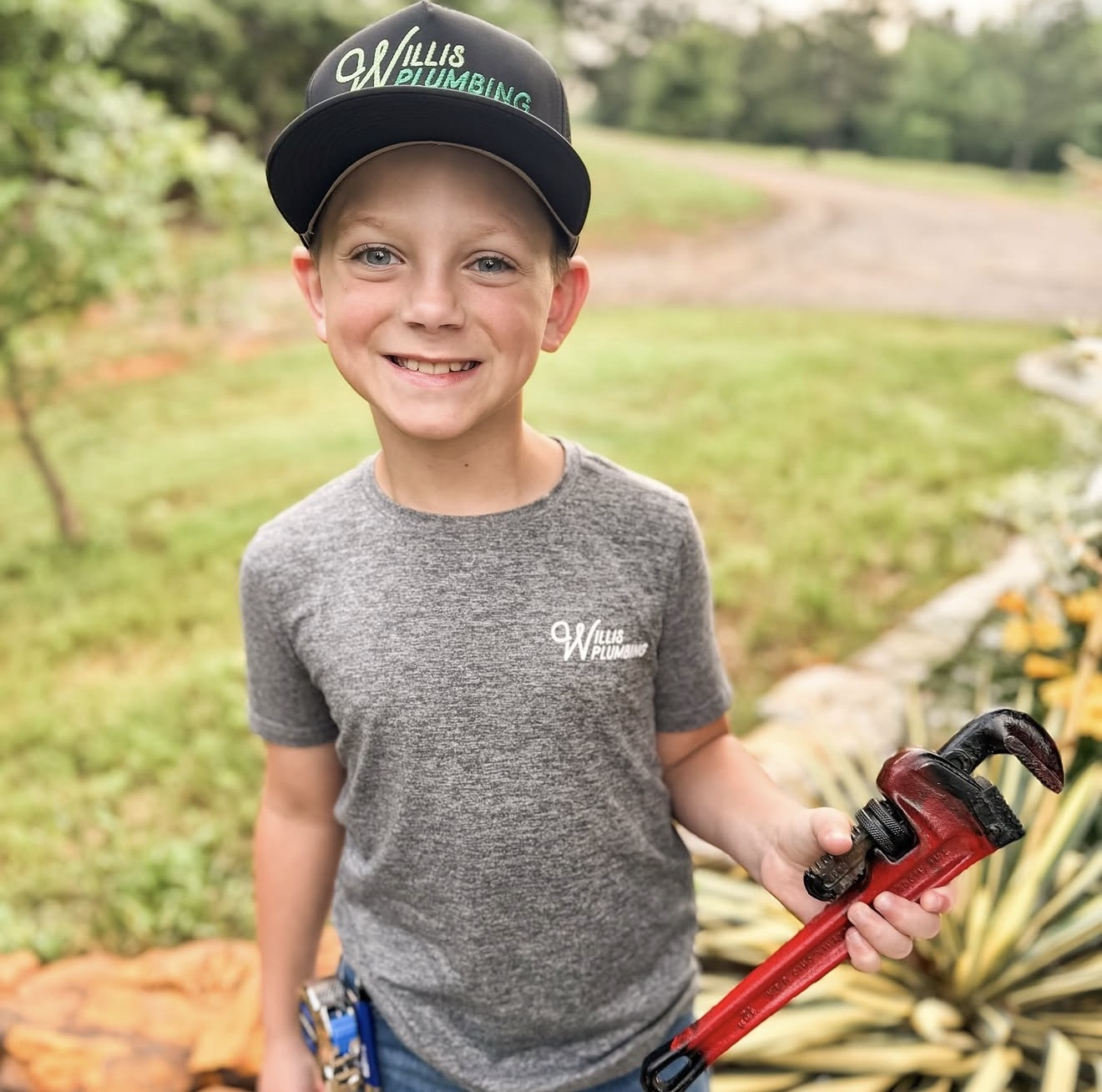A young boy smiling outdoors, wearing a black cap and gray t-shirt with 'Willis Plumbing' logo, holding a red pipe wrench.