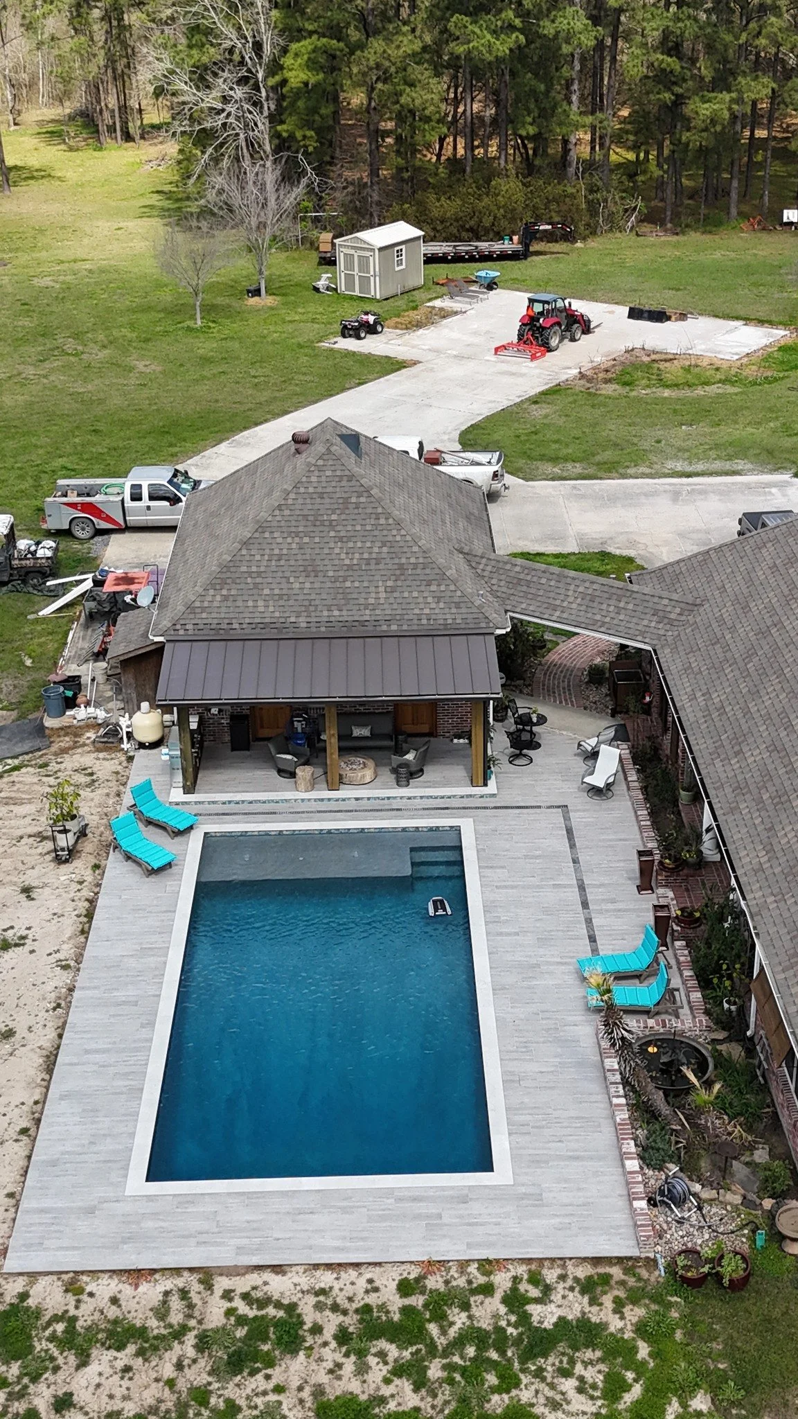 An aerial view of a backyard with a swimming pool, patio furniture, a house with a porch, and a large driveway with vehicles and yard equipment. There is a wooded area in the background.