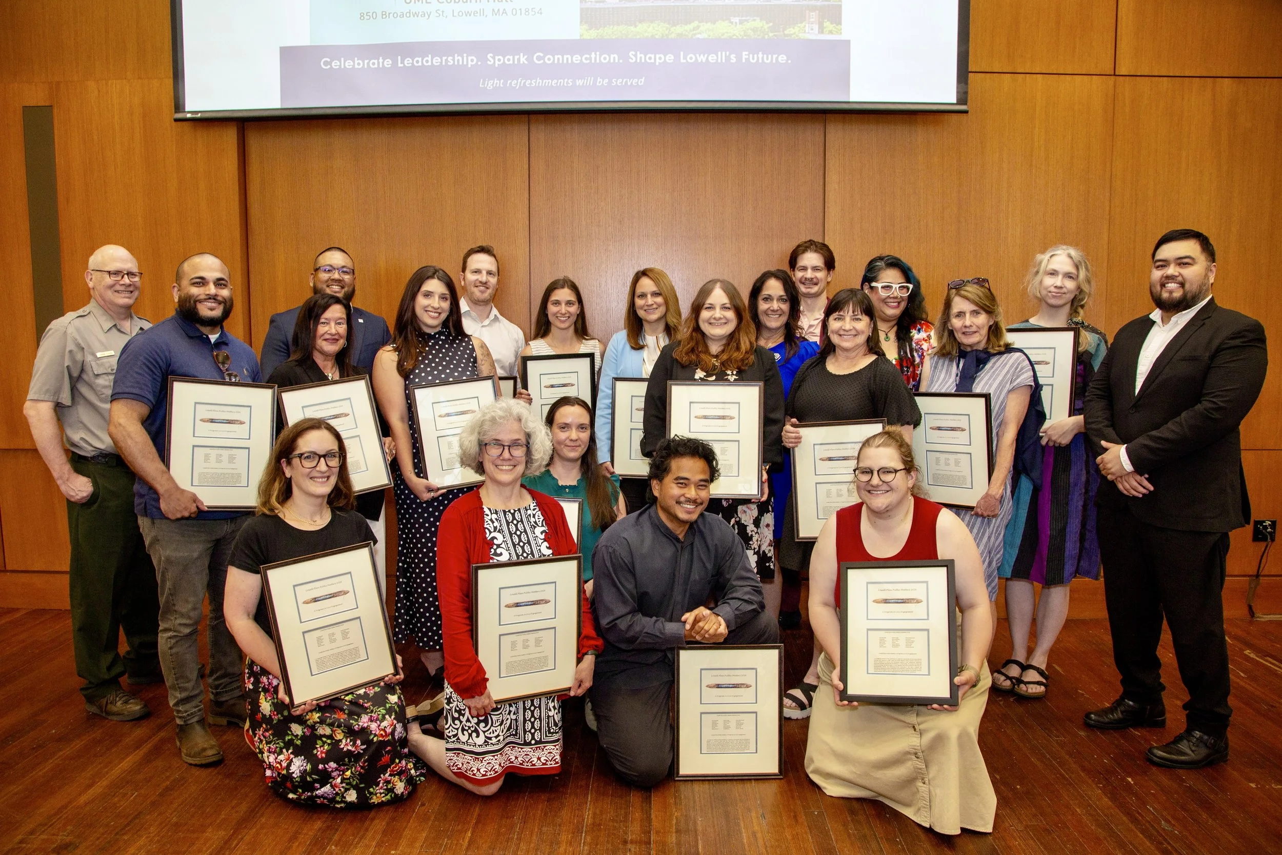 Group of diverse people smiling and holding framed certificates in a room with wooden walls. A presentation screen is visible above them.