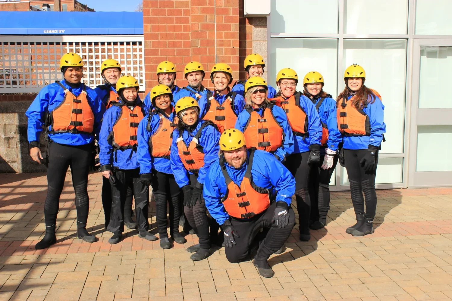 Group of people wearing yellow helmets, orange life jackets, blue jackets, and black gloves posing outdoors on a brick pavement, in front of a building with large windows and a brick wall.