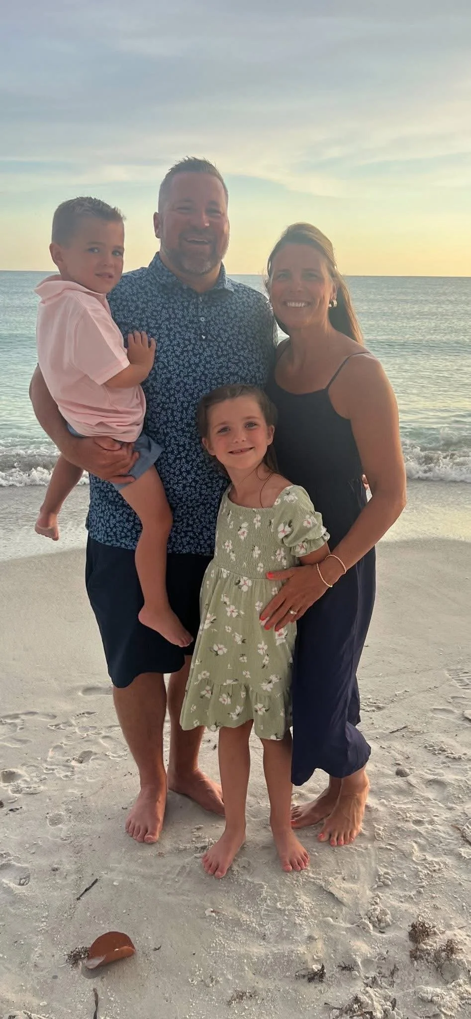 A family of four, including a man, woman, and two children, standing barefoot on the beach with the ocean and sunset in the background.