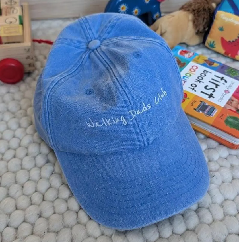 Blaue Baseballkappe mit aufgesticktem Schriftzug 'Walking Dad's Club' auf einem weißen, gepunkteten Teppich, im Hintergrund Spielzeug und Zeitschriften.