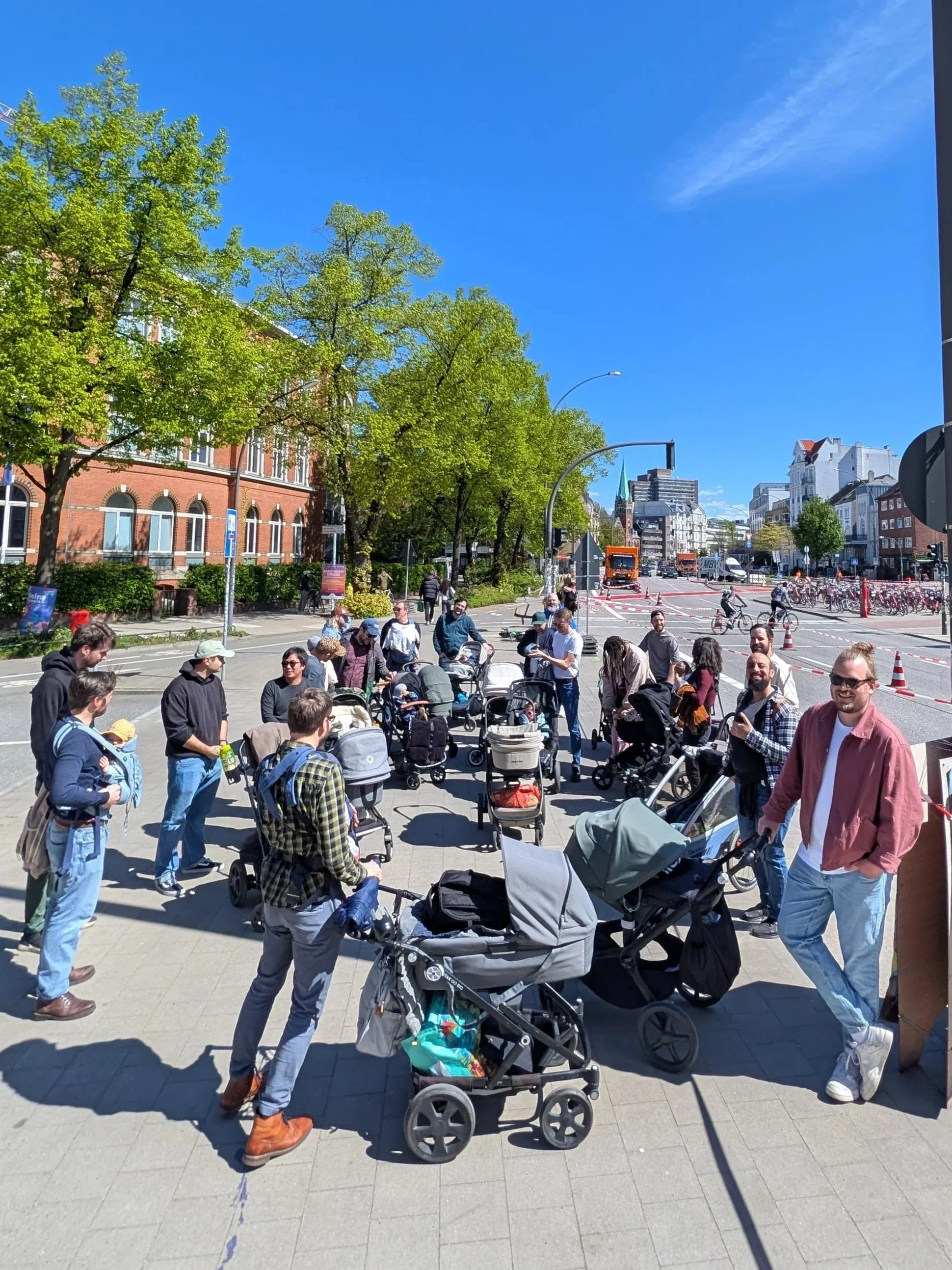 Menschen versammeln sich mit Kinderwagen auf einer Stadtstraße bei schönem Wetter, im Hintergrund sind Bäume, Gebäude und Fahrräder zu sehen.