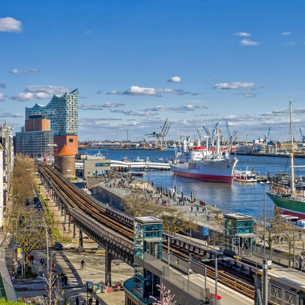 Ein Hafen mit mehreren Schiffen und Booten, moderner Architektur im Hintergrund, Eisenbahnlinien im Vordergrund, bei sonnigem Himmel.