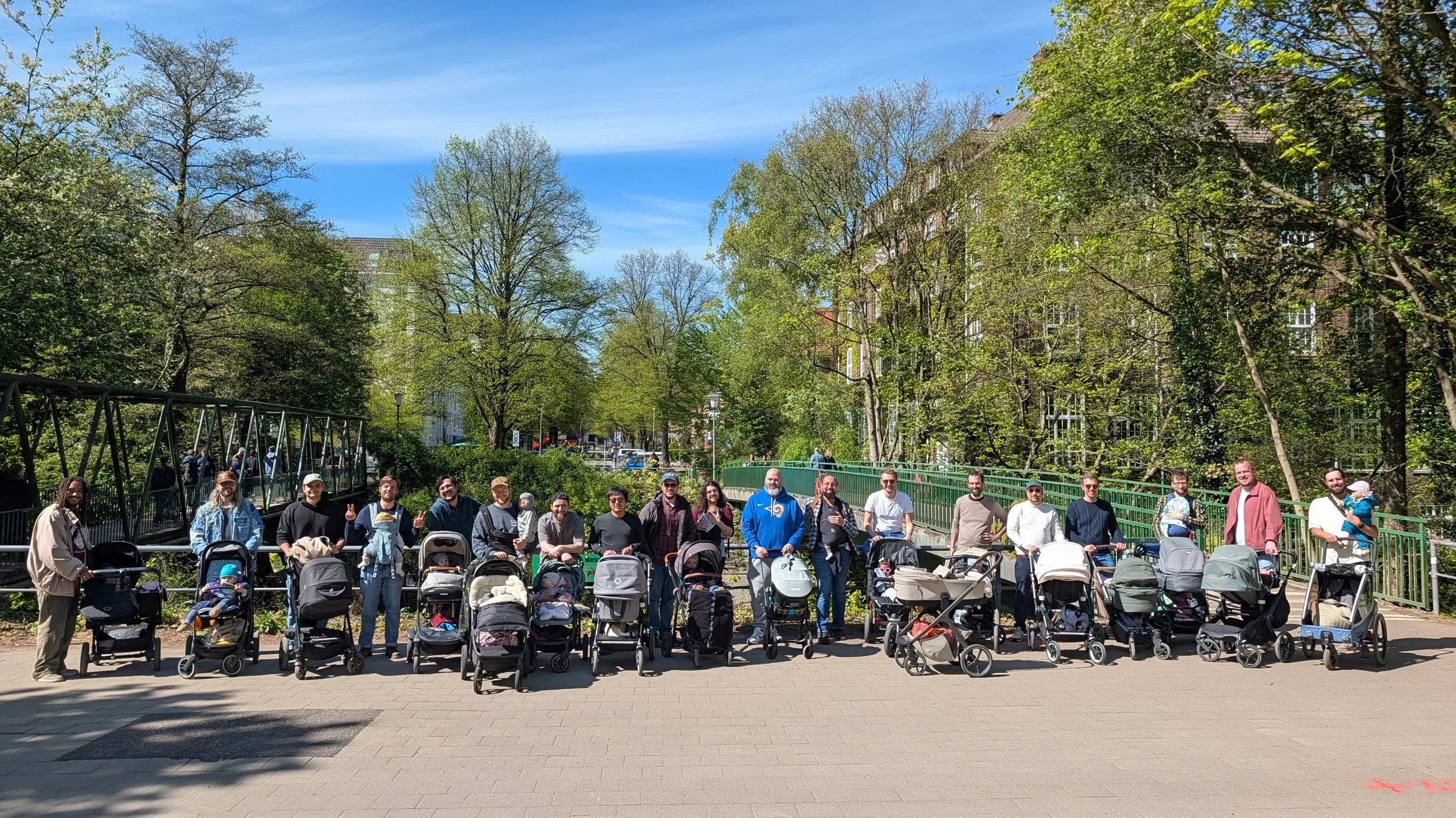 Eine Gruppe von Menschen, darunter viele Eltern mit Kinderwagen, versammelt sich im Freien in einem Park mit grünen Bäumen und Gebäuden im Hintergrund an einem sonnigen Tag.