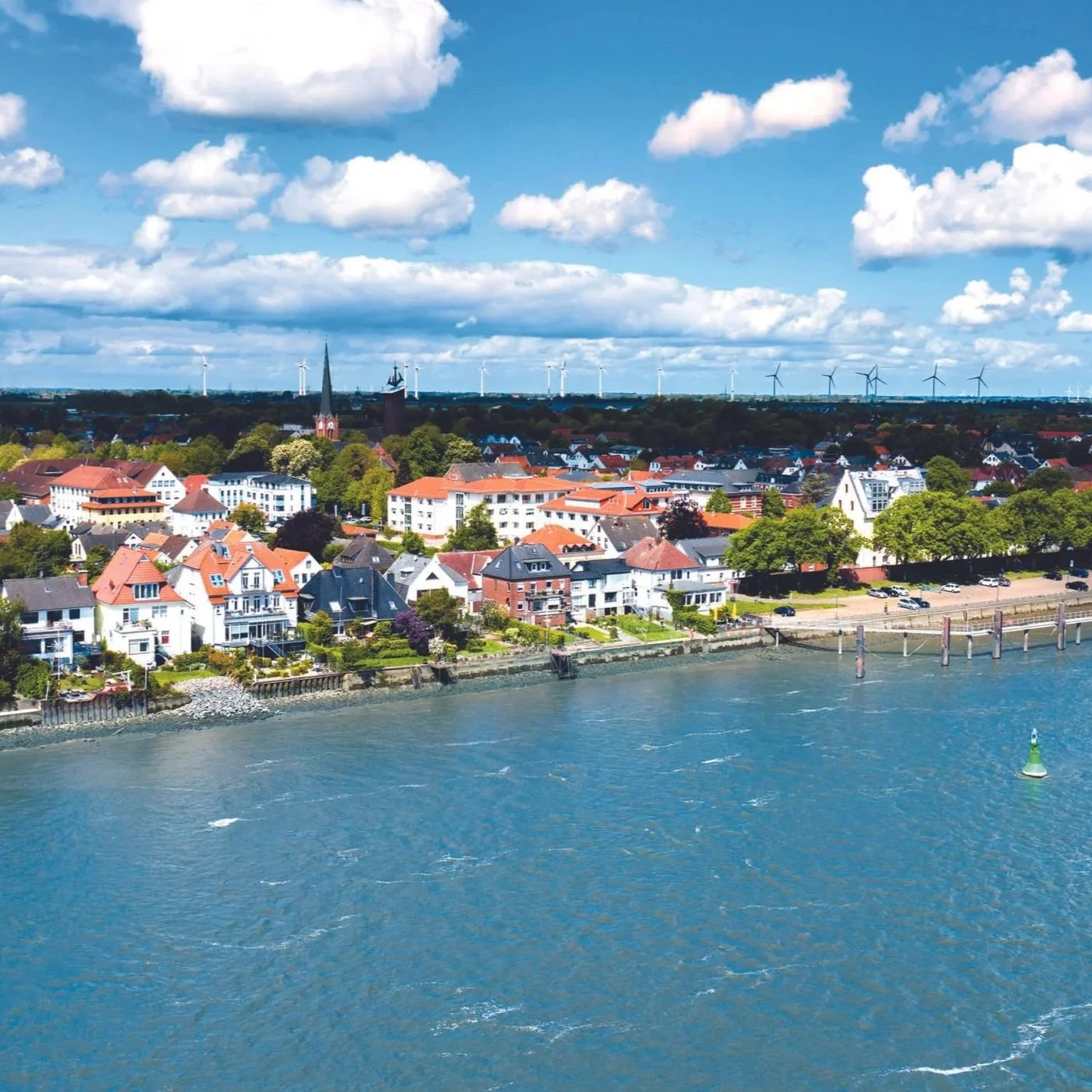 Luftaufnahme einer Stadt am Fluss mit Wohnhäusern, Kirchen und Windkraftanlagen im Hintergrund bei blauem Himmel mit Wolken.