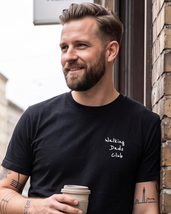 Ein lächelnder Mann mit Bart und kurzen Haaren, trägt ein schwarzes T-Shirt mit weißem Aufdruck "Walking Dads Club". Er hält einen To-Go-Kaffee und steht neben einer Ziegelmauer, im Hintergrund ein Gebäude.