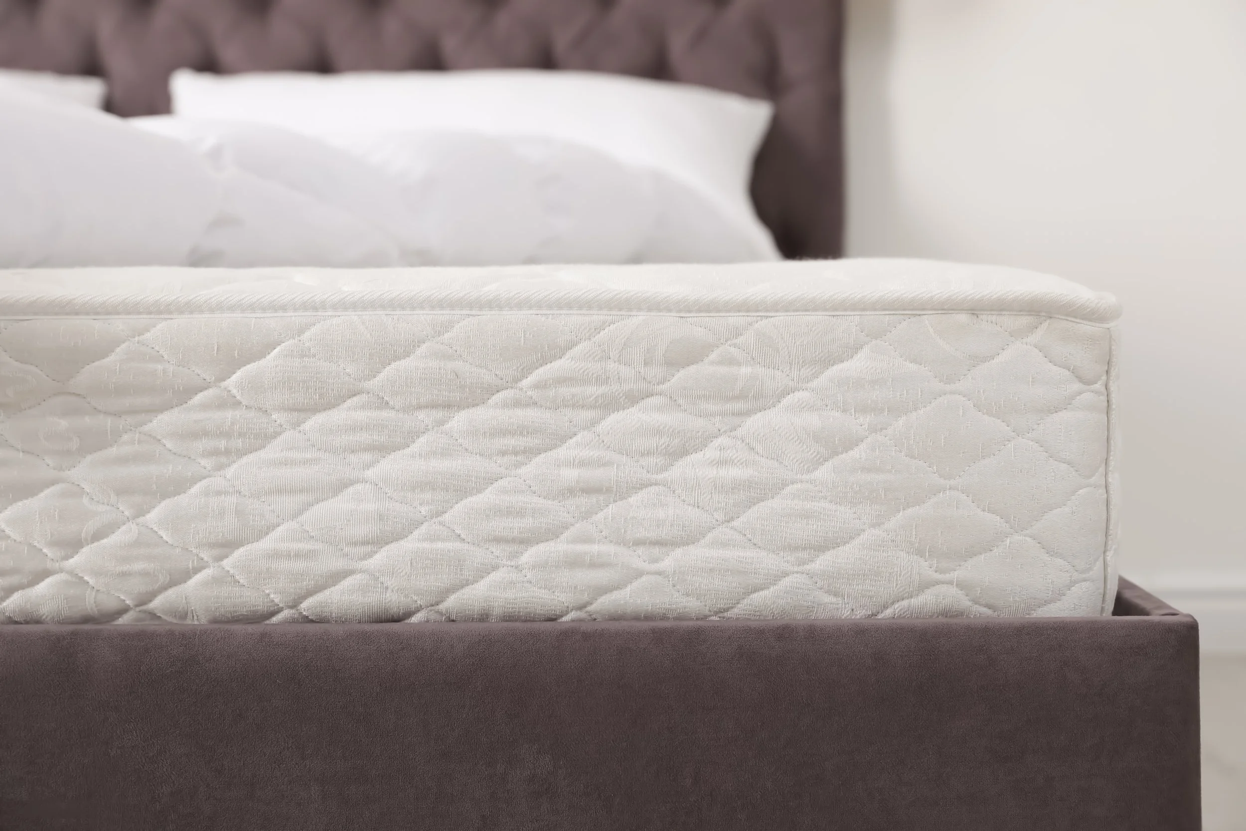 Close-up of a mattress with a quilted pattern on a dark bed frame with pillows in the background. This represents a safe place to sleep.