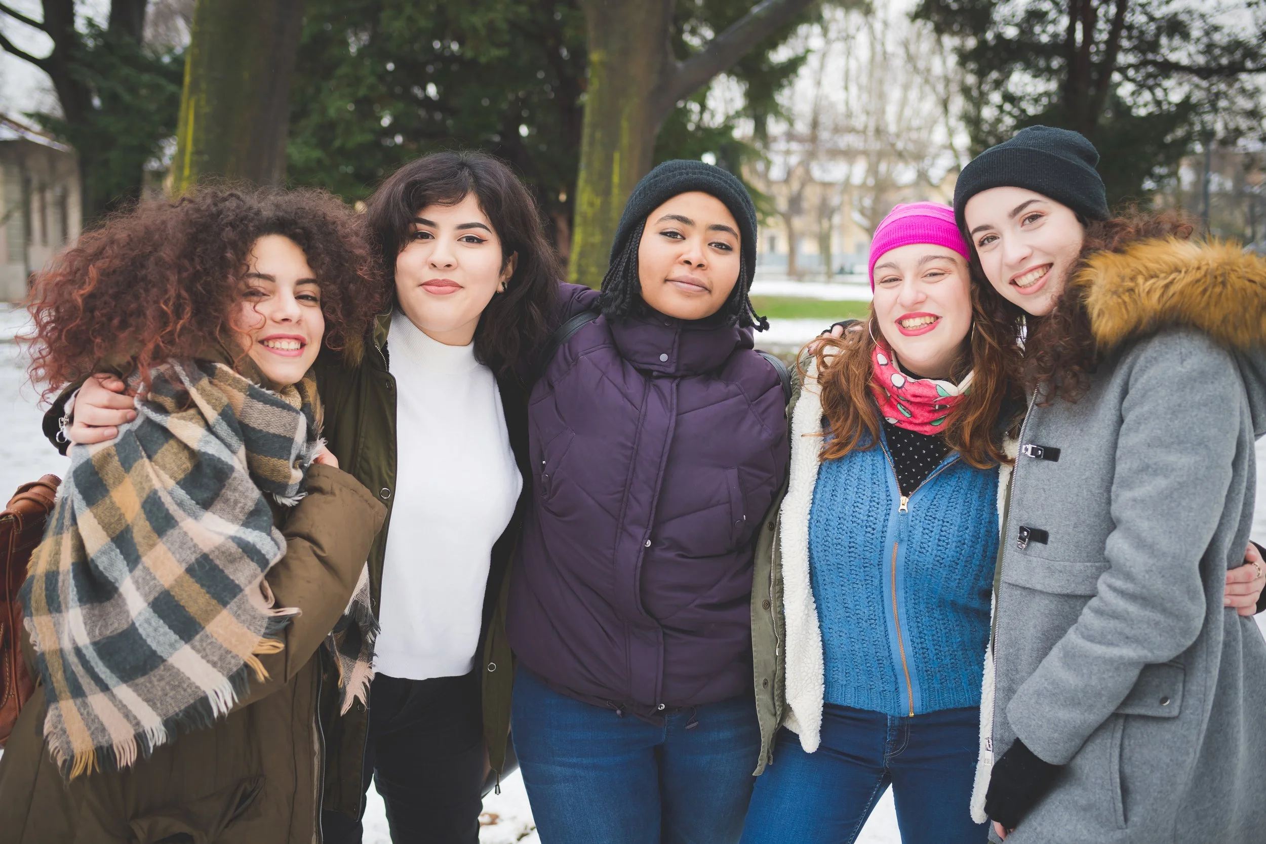 Group of six women standing close together outdoors on a cloudy day, dressed warmly with winter hats and coats, with trees and buildings in the background.