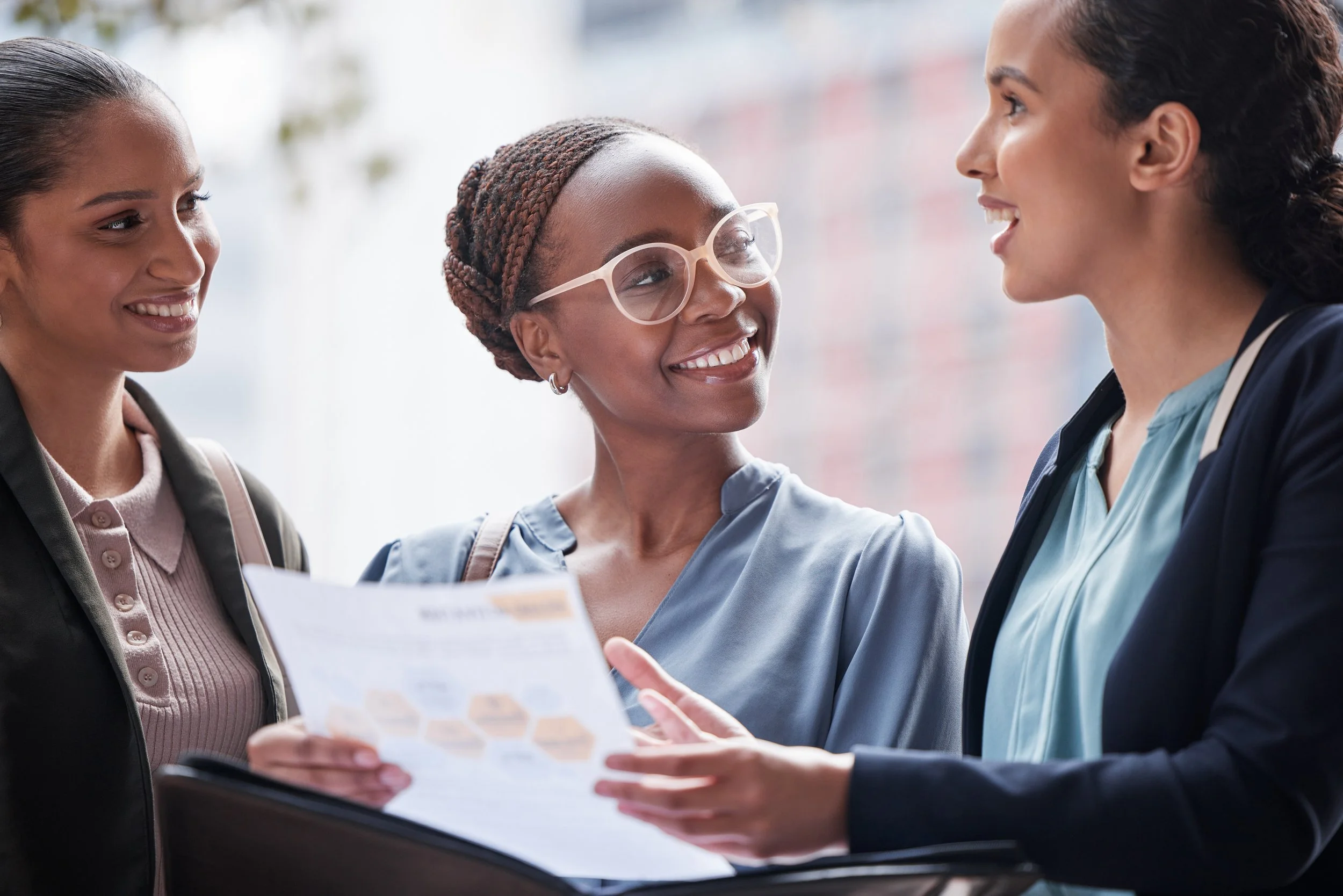 Three women, two with dark hair and one with braided hair and glasses, smiling and talking, with one holding a clipboard or document.