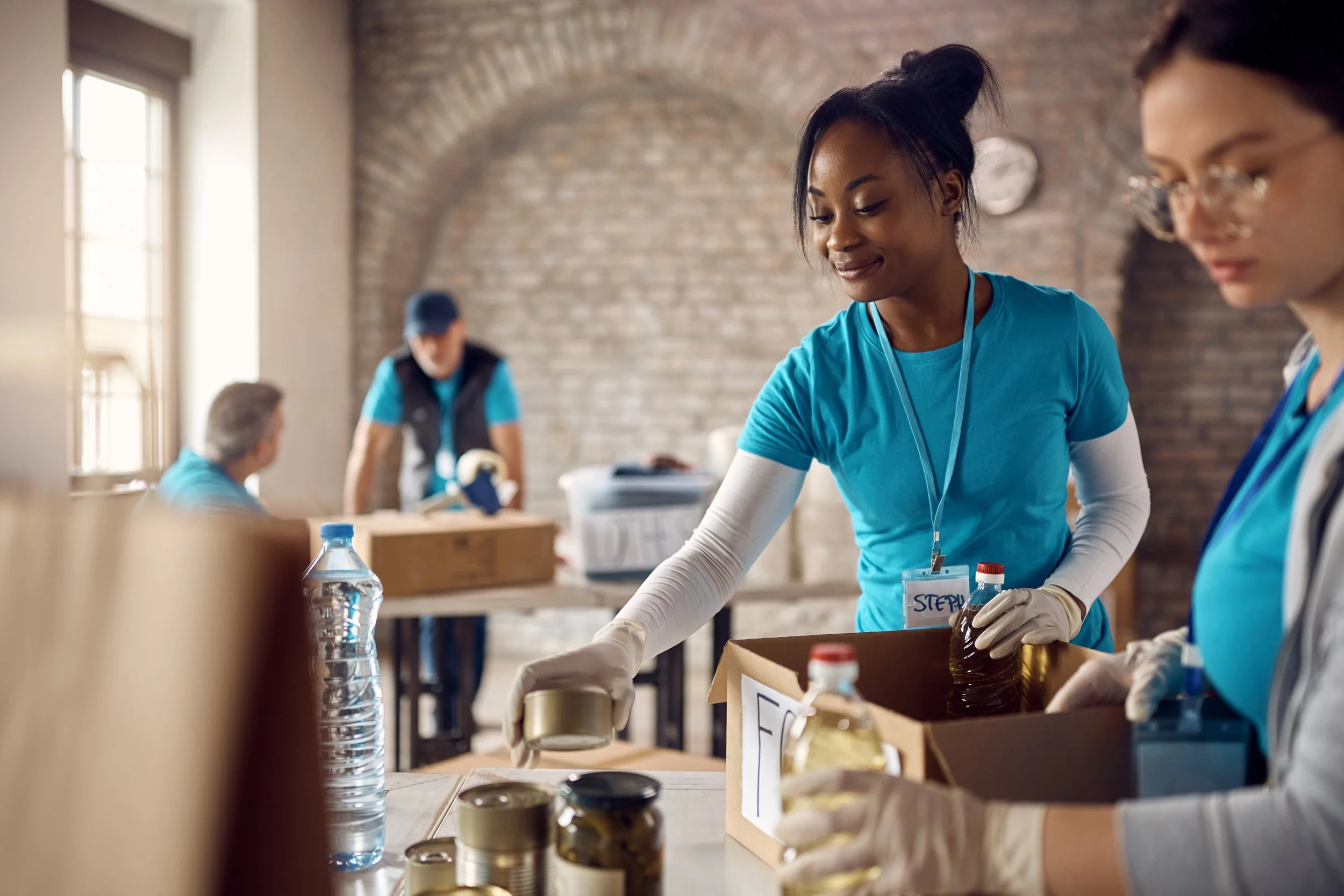 Two women volunteers working together at a community service event, packing supplies in a room with large windows and brick walls.