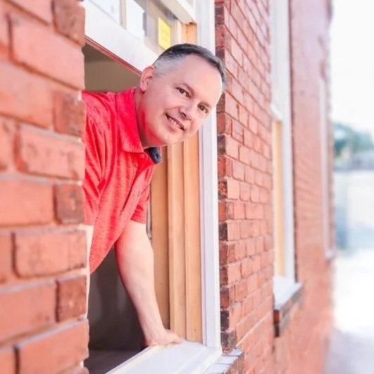 A man in a red shirt leaning out of a second-story window of a brick building, smiling at the camera.