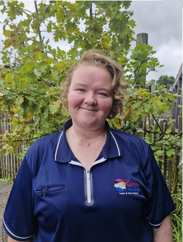 A smiling woman standing outdoors in front of green leafy plants, wearing a navy blue polo shirt with a logo that says "Karinga Children's Learning & Care Nursery."