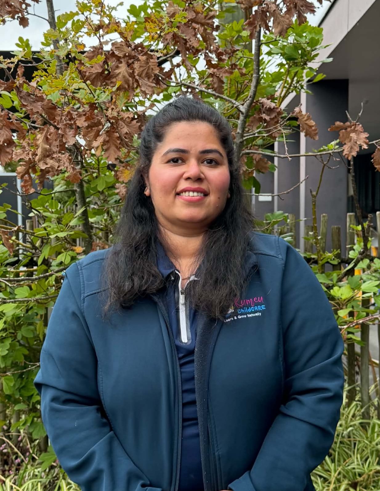 A woman with long dark hair smiling outdoors in front of a tree with brown and green leaves, wearing a navy blue jacket with a childcare logo.