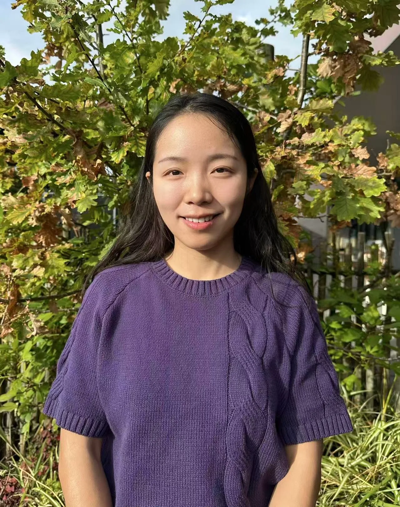A young woman with long black hair smiling outdoors, wearing a purple sweater, with green and brown leaves and trees behind her.