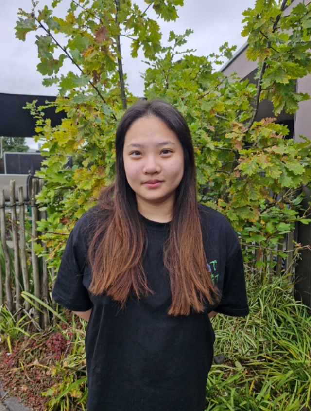 Young woman with long brown hair standing outdoors in front of a green leafy shrub, wearing a black T-shirt.