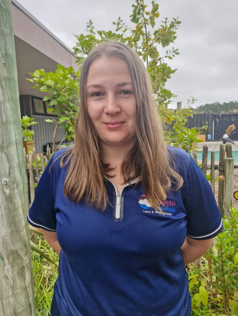 Young woman with long brown hair wearing a navy blue polo shirt outdoors in a garden area with plants and trees under a cloudy sky.