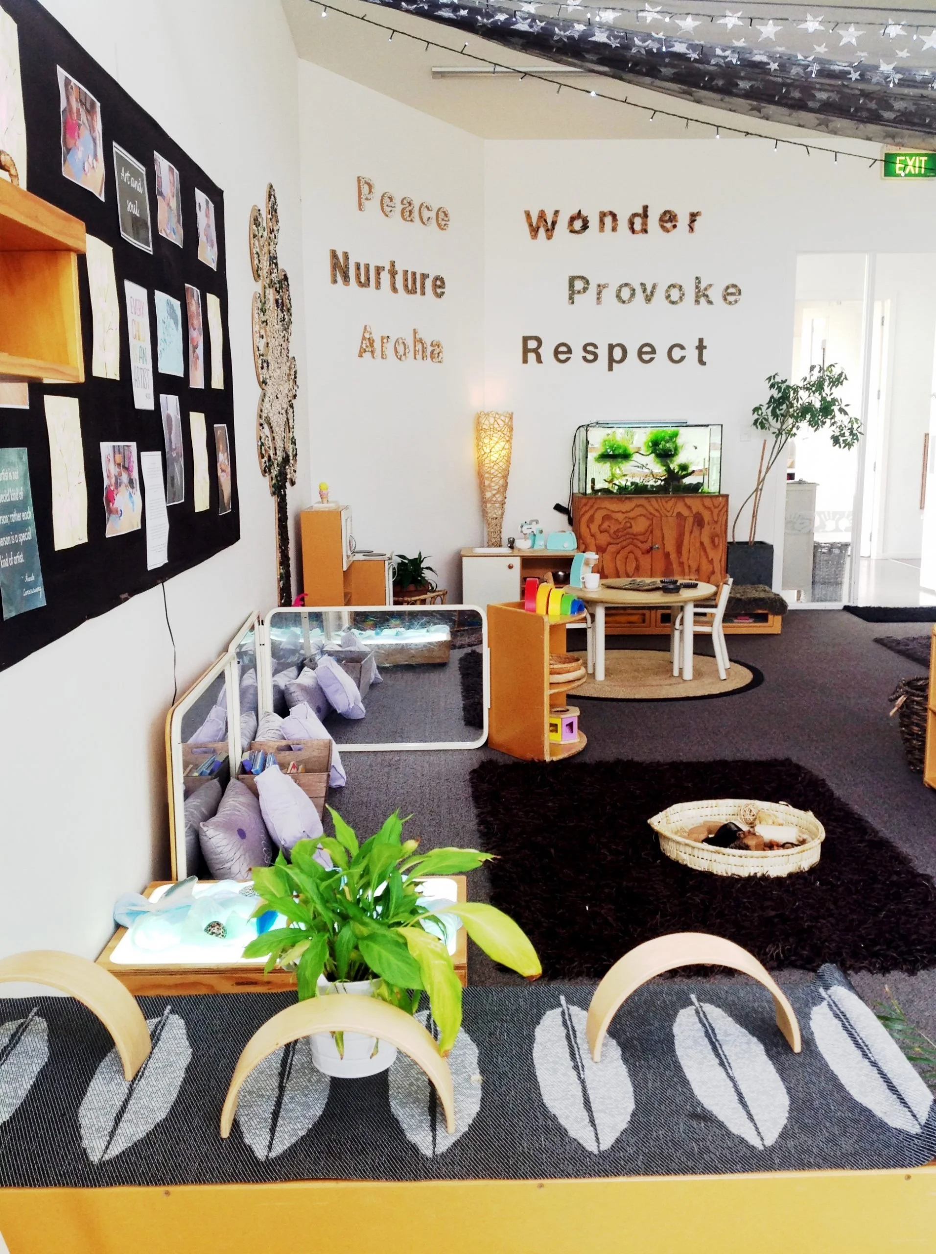 A cozy indoor space with a black and white striped rug, a round table with wooden chairs, a green potted plant, black shaggy rug, a children's play area with pillows, a small fish tank, and colorful lamp, wall decorations with words like 'Peace,' 'Nurture,' 'Aloha,' 'Wonder,' 'Provoke,' 'Respect,' and photos on black display board, along with a tall light stand and a door leading outside.