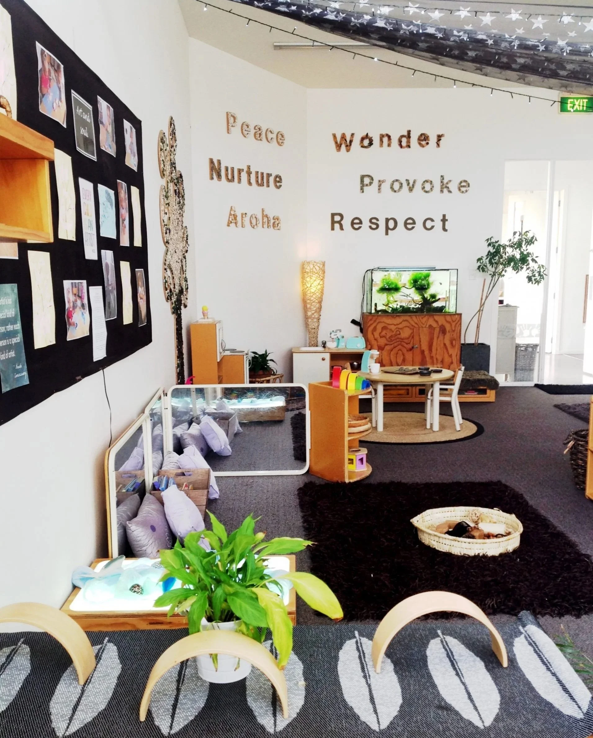 Interior of a room decorated with motivational and cultural words on the wall, a black display board with photos and papers, a small table with toys, an artificial fish tank, lush green plants, a round black rug, wicker basket, and chairs with light wood frames. The ceiling has star-patterned drapery and string lights, and there is an exit sign near the door.