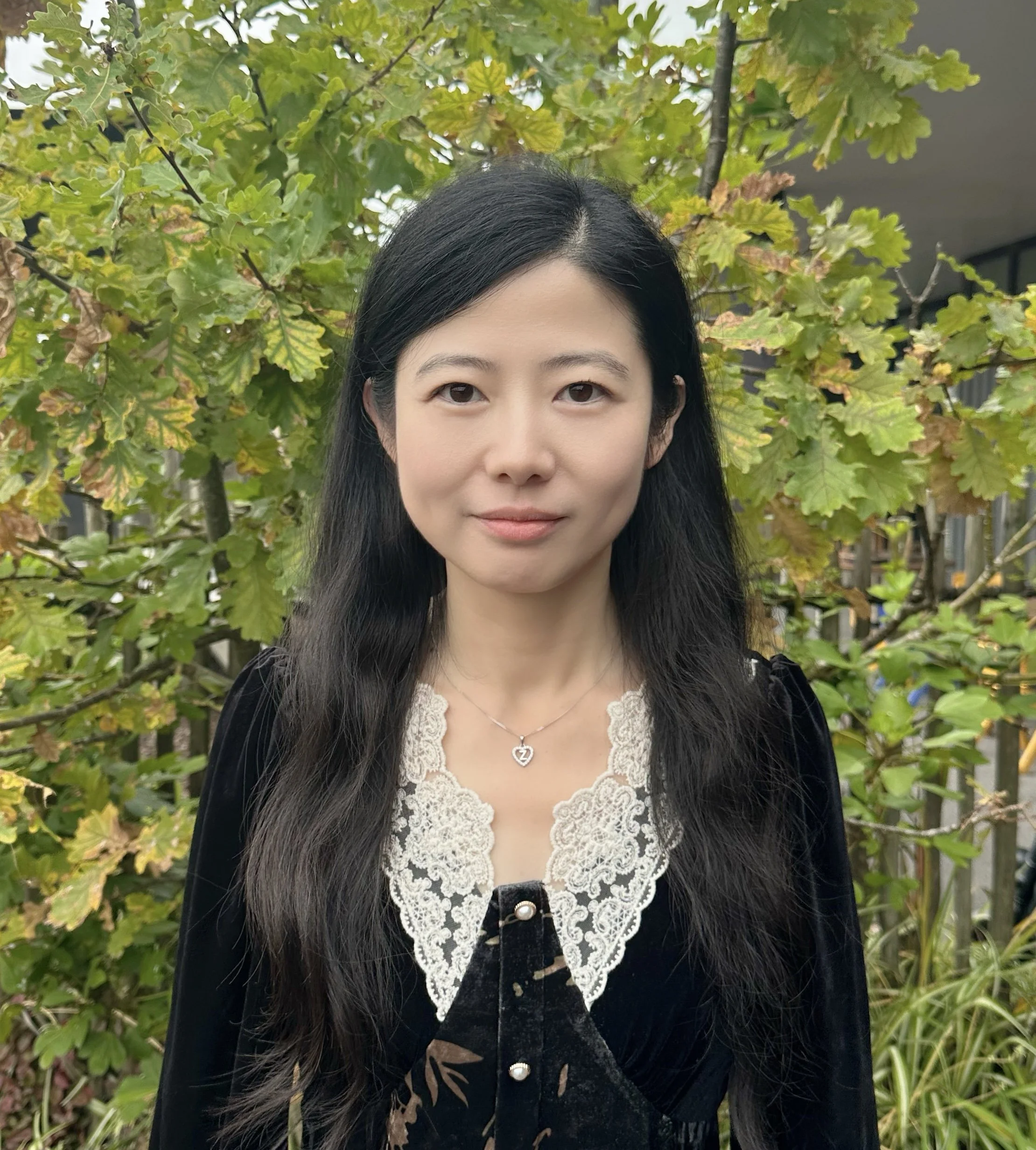 A woman with long black hair standing outdoors in front of a leafy green tree, wearing a black and white lace top and a necklace with a heart-shaped pendant.