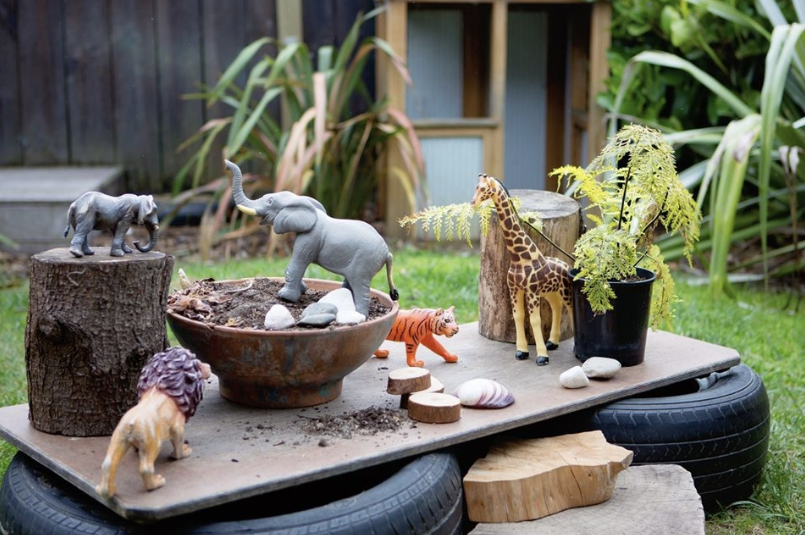 Children's toys placed on a homemade table made from tires and wooden planks in a garden, including toy animals like an elephant, lion, tiger, giraffe, and elephants, with plants and greenery in the background.