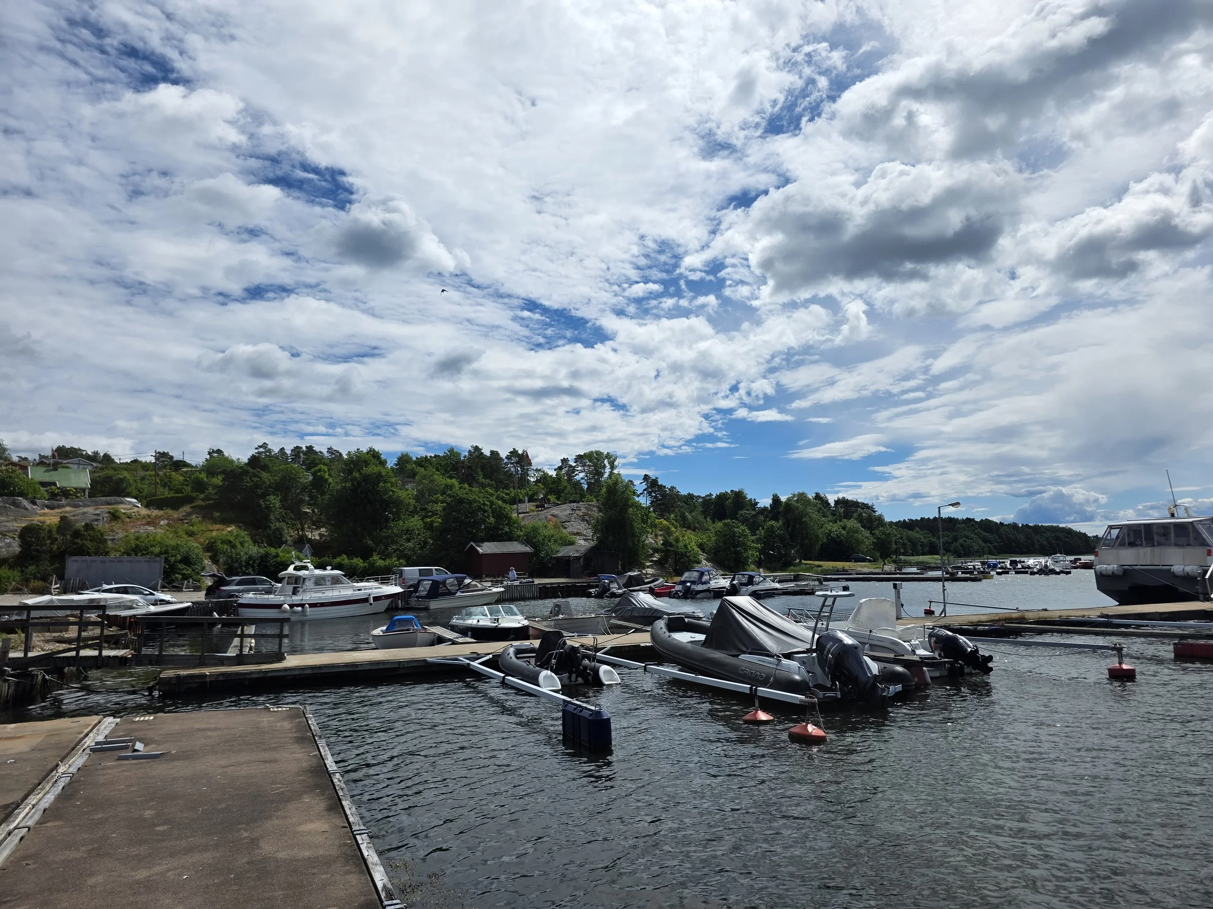 En marina med båter og speedbåter som er fortøyd, omgitt av grøntområder og hus under en delvis skyet himmel.