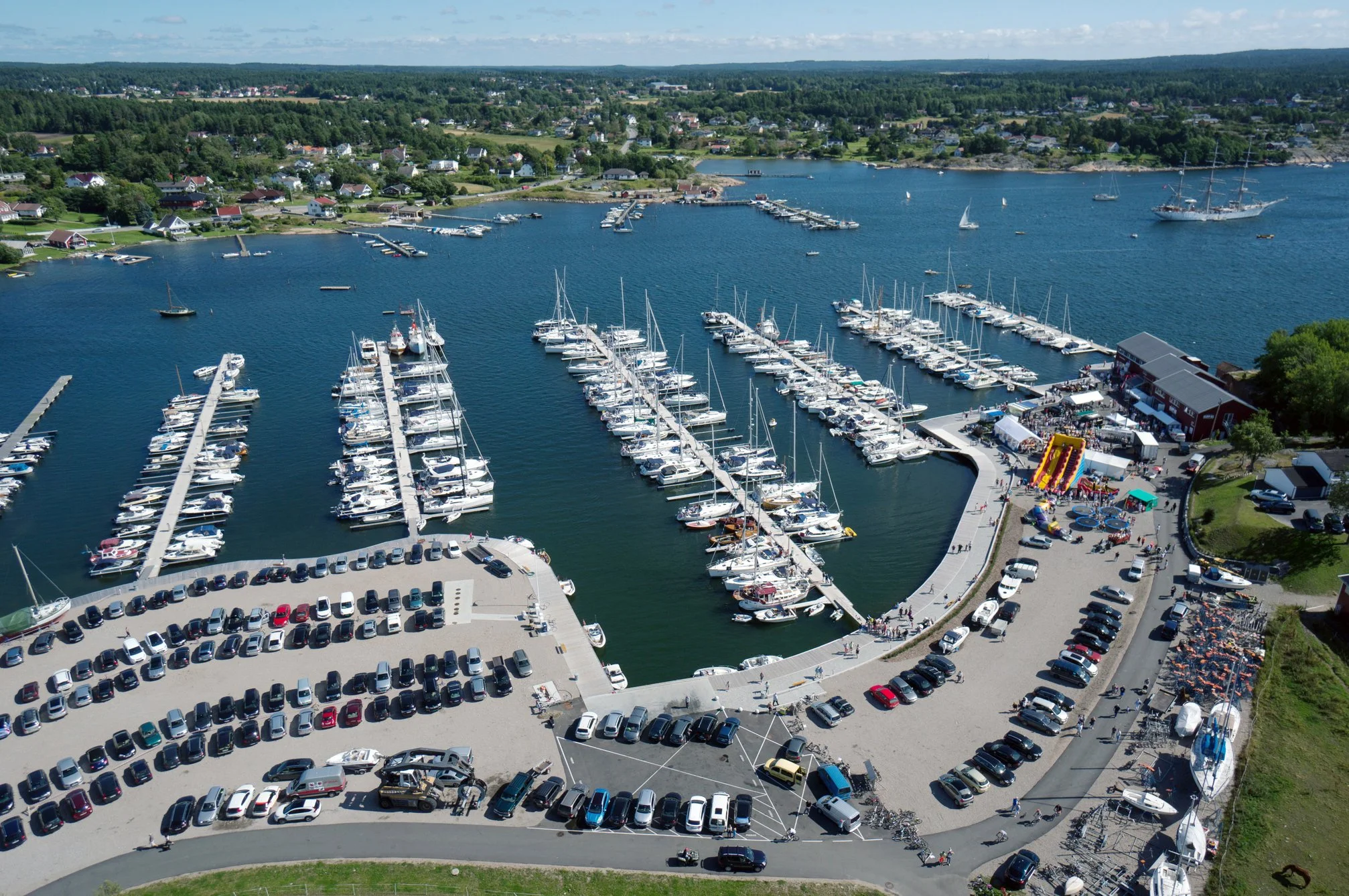En flyfoto av en liten marina med mange båter og en parkeringsplass med biler. Det er også folk som går rundt og en forlystelse med sklier i nærheten.