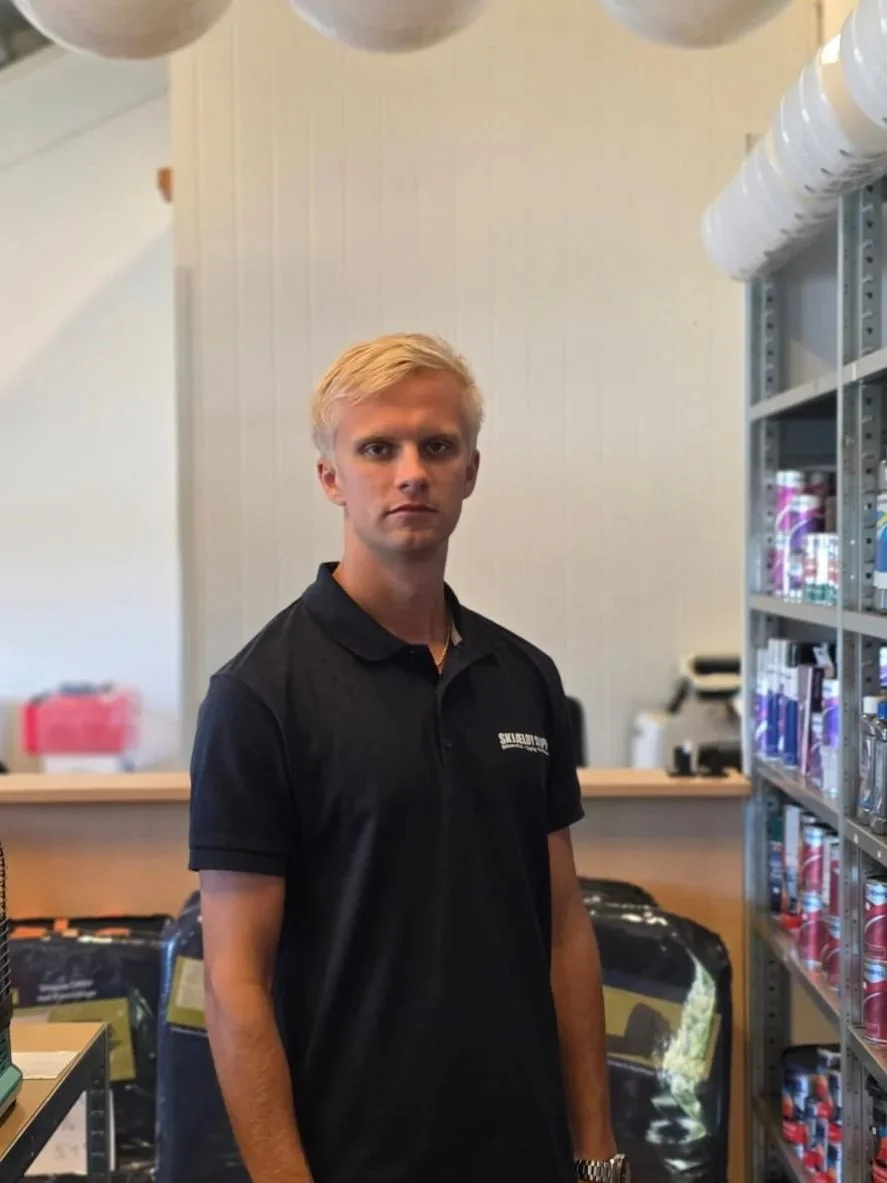 En mann med blekt, blond hår og svarte klær står inne i en butikk, med hyller fylt med ulike produkter. Han ser direkte mot kamera.