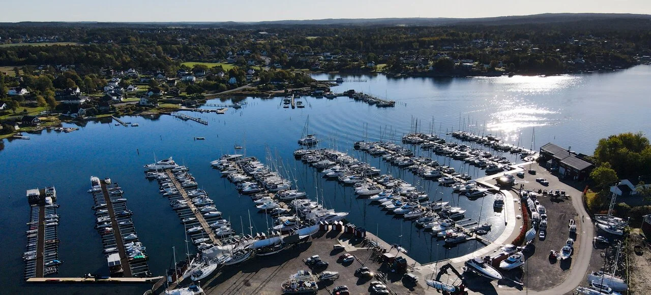 Fjernbilde av en marina med mange båter og sjøfartøy, omgitt av boligområder og natur.