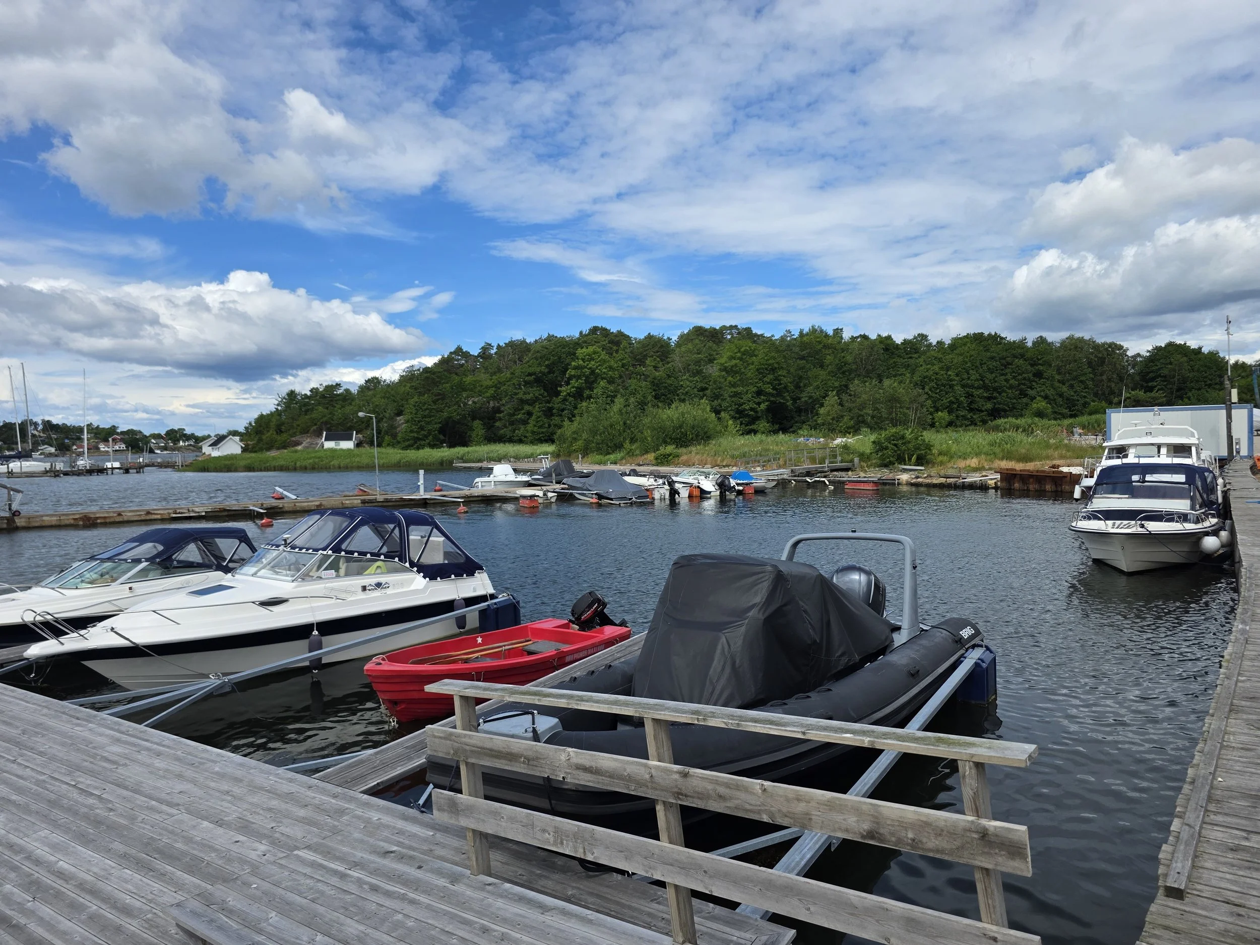 Båter og robåter som er fortøyd ved en kai med en grønn ås og en blå himmel med skyer.