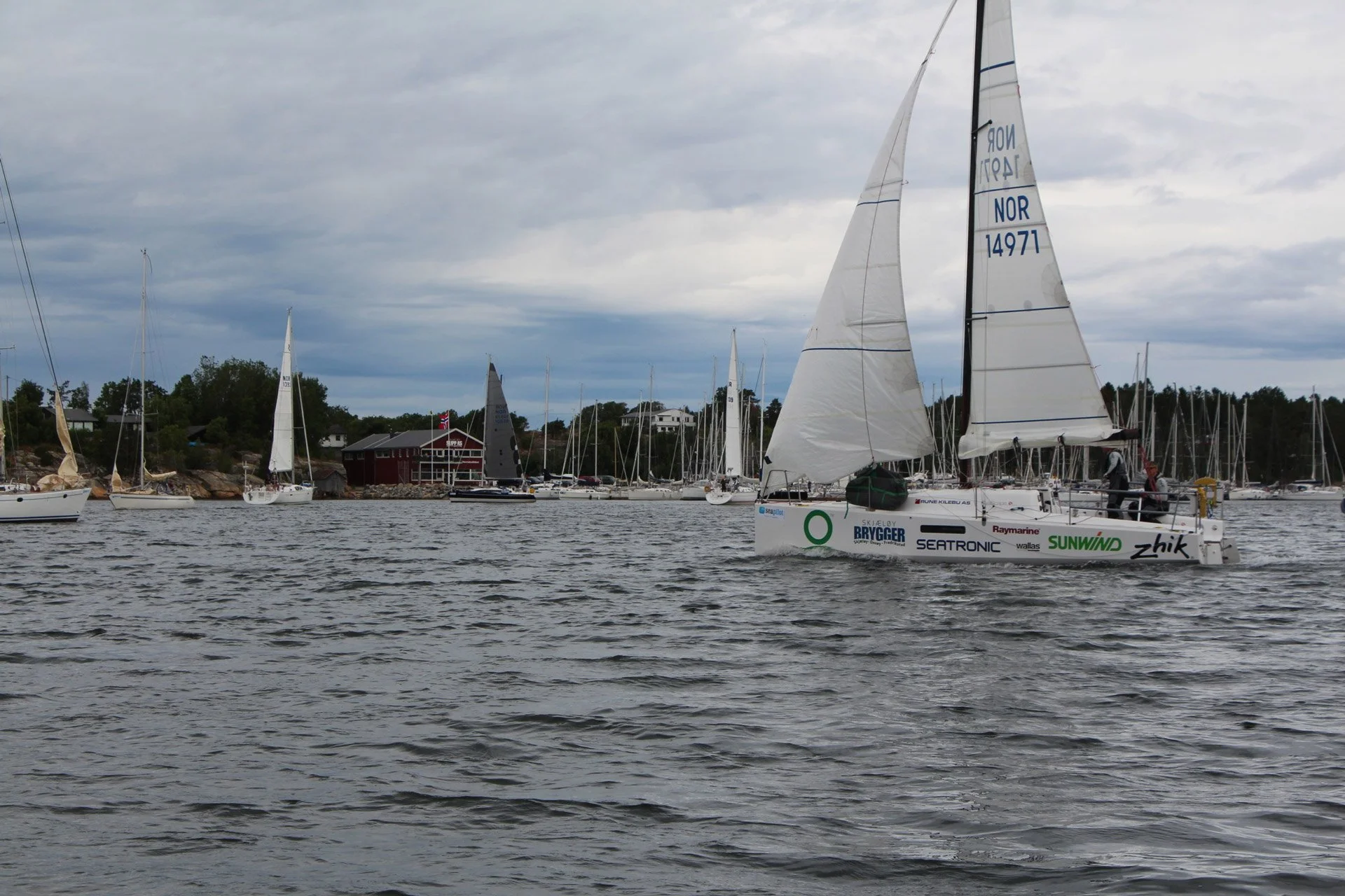 Sailbåt på vannet med flere andre båter og en bygning i bakgrunnen. Overskyet vær.