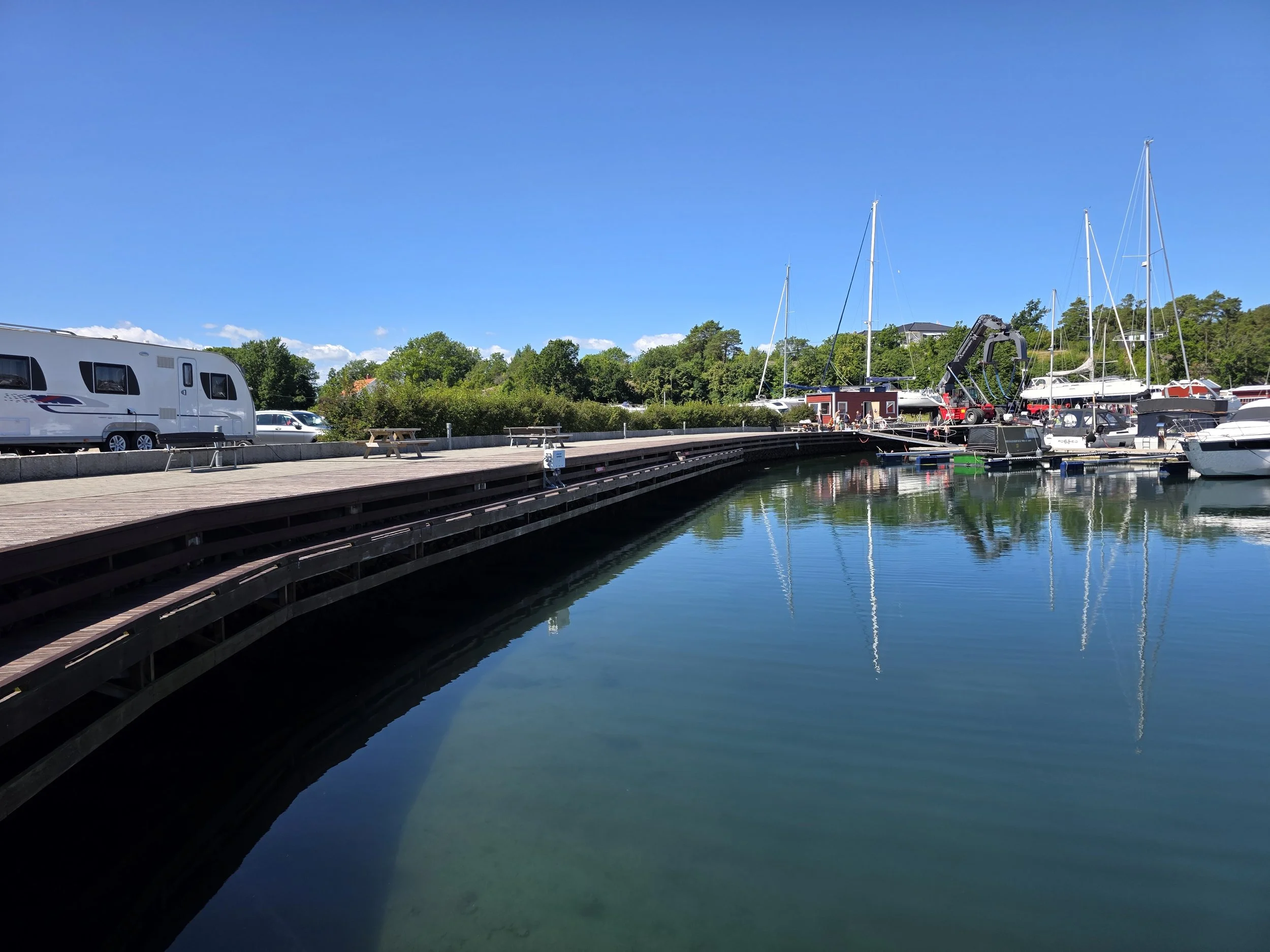 Båter og seilbåter i en marina med blå himmel og grønne trær i bakgrunnen.