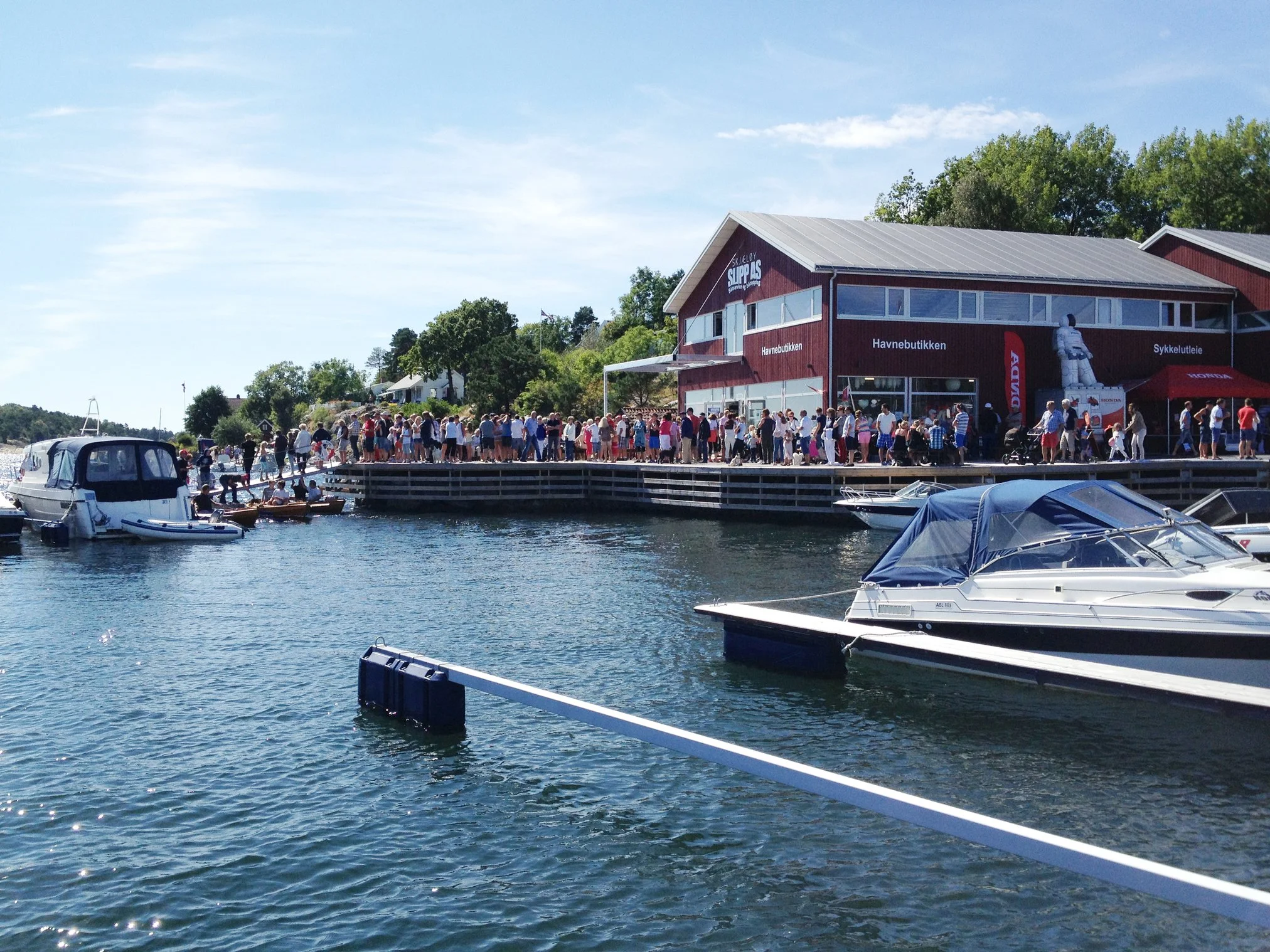 En gruppe mennesker står på en kai ved vannet, med båter i vannet foran og en rød bygning med store vinduer bak. Menneskene ser ut til å delta i en fest eller arrangement.