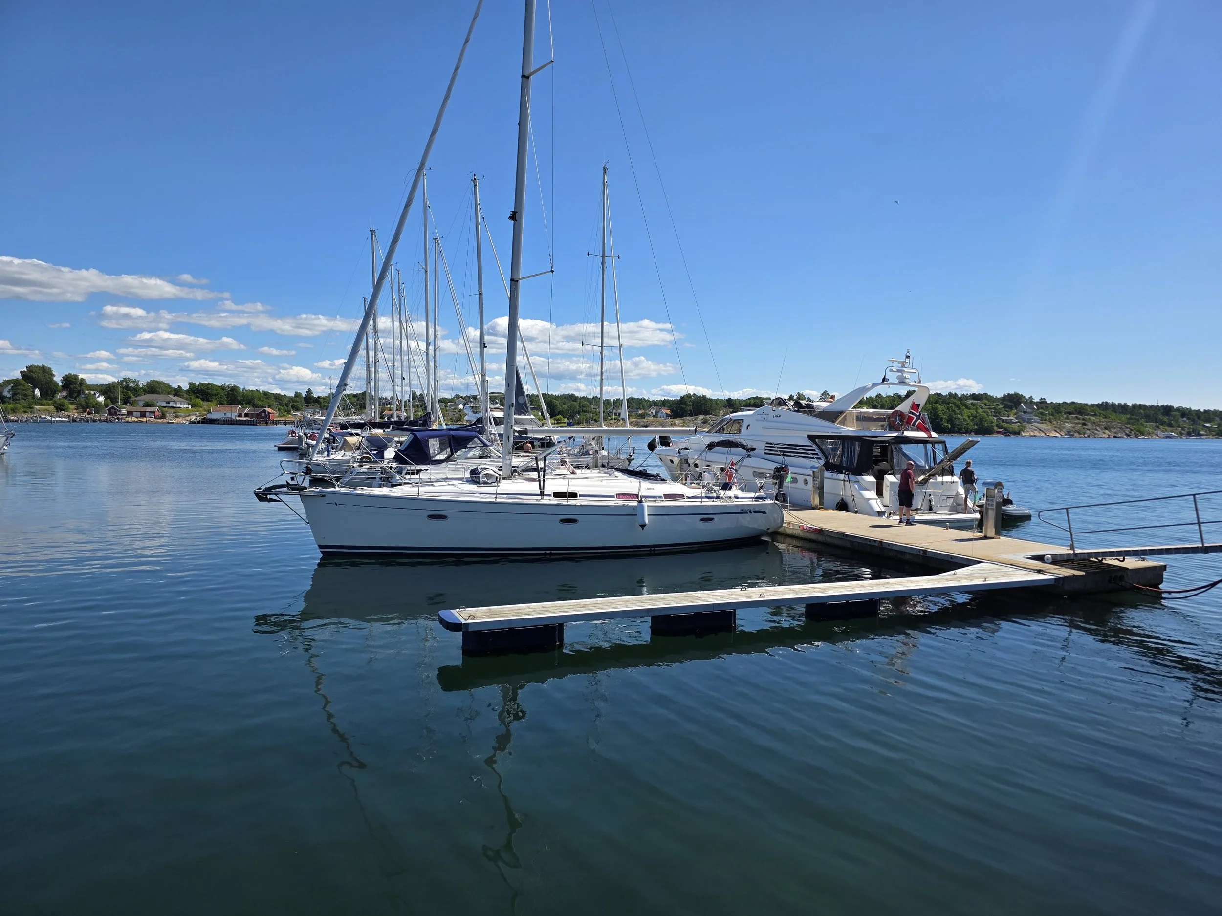 Båter og yacht ligger ved en kai på en klar, solrik dag med blå himmel og noen skyer.