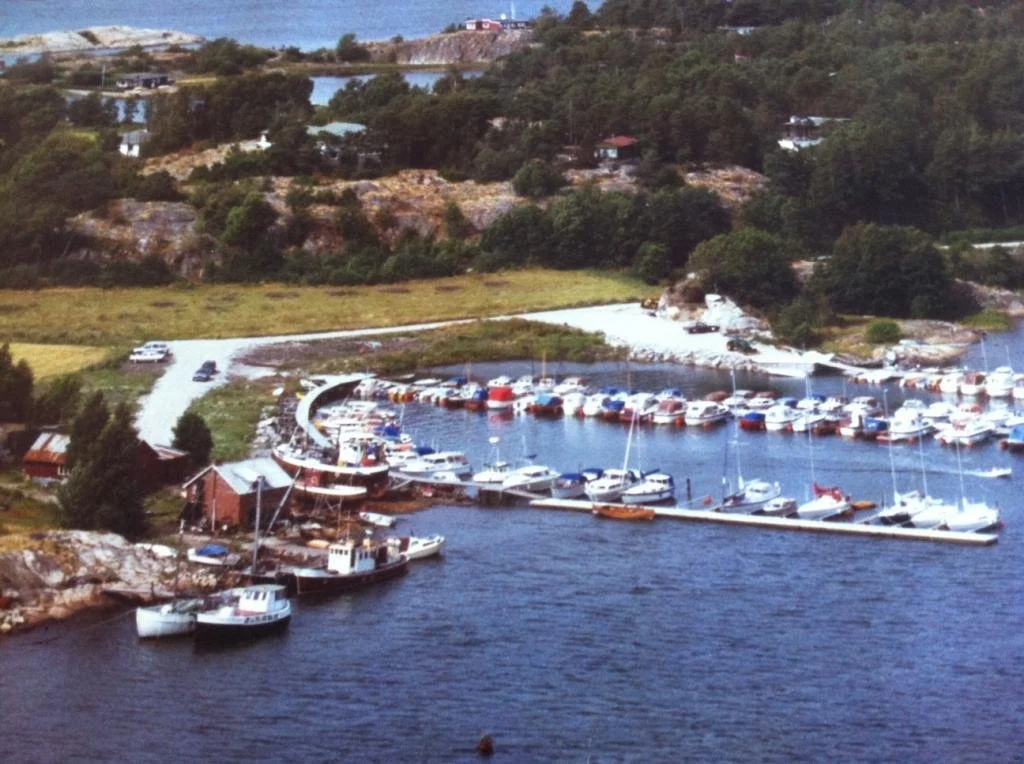 En liten båthavn med mange båter og en vei som går rundt havnen, omgitt av grønne åser og hus i bakgrunnen.