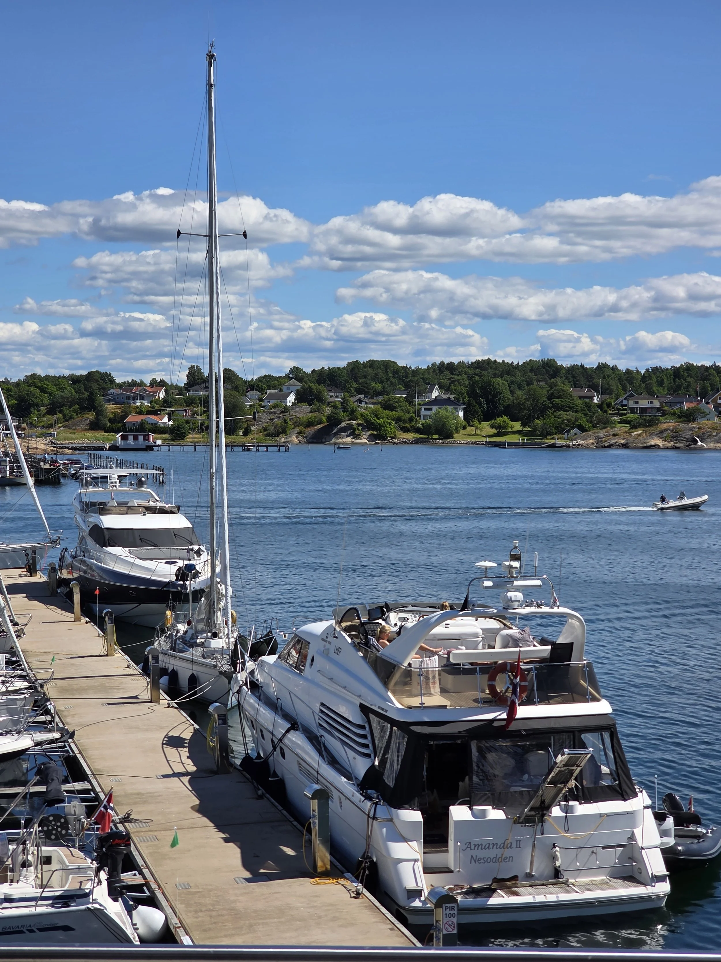 Båter og yachts i en havn med hus og grønne trær i bakgrunnen under en blå himmel med spredte skyer.