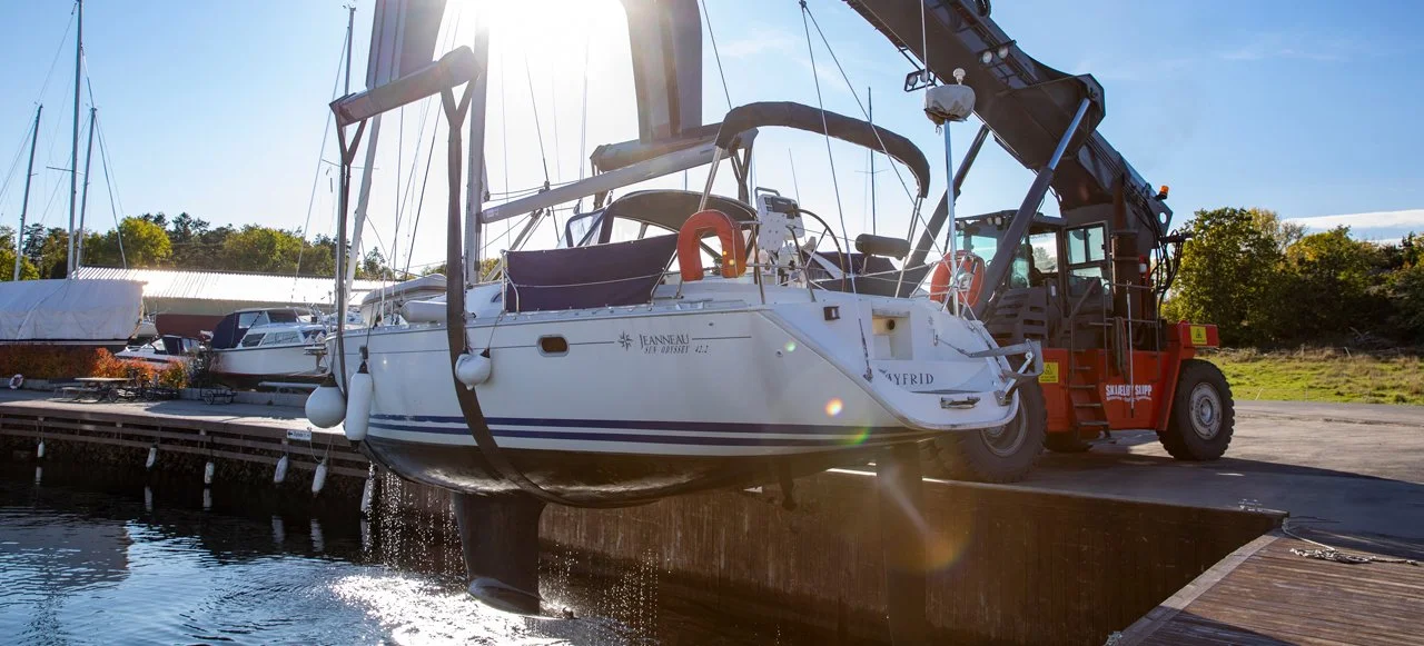 En hvit seilbåt blir hevet opp av en kran ved en marina, med flere andre båter i bakgrunnen og klar blå himmel.
