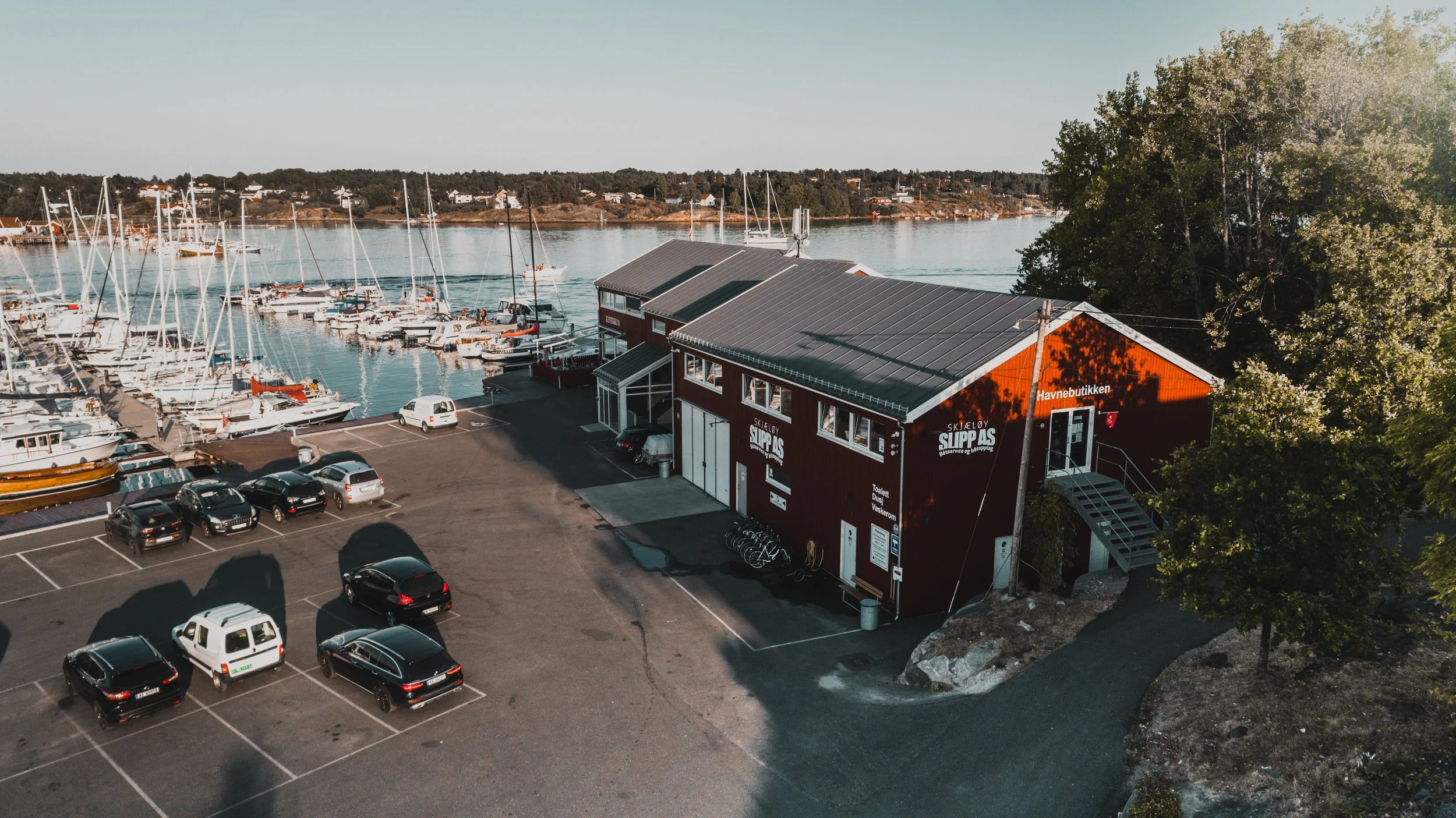 Båt- og yacht-havn med biler parkert foran en rød bygning, omgitt av vann og trær.