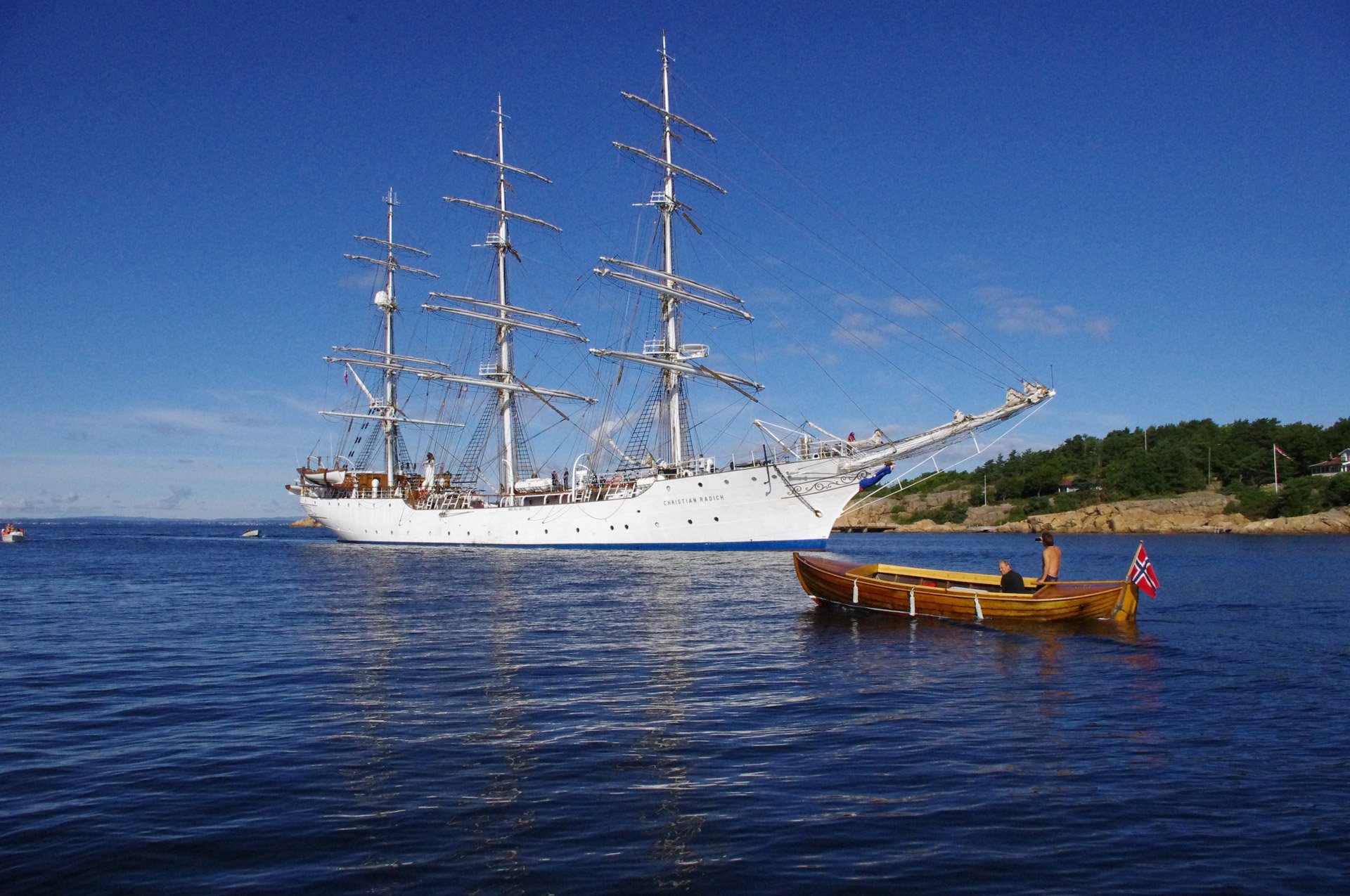 Et stort hvitt seilskip med tre master i vannet, en liten robåt med to personer og et norsk flagg på den, og en kystlinje med trær i bakgrunnen.
