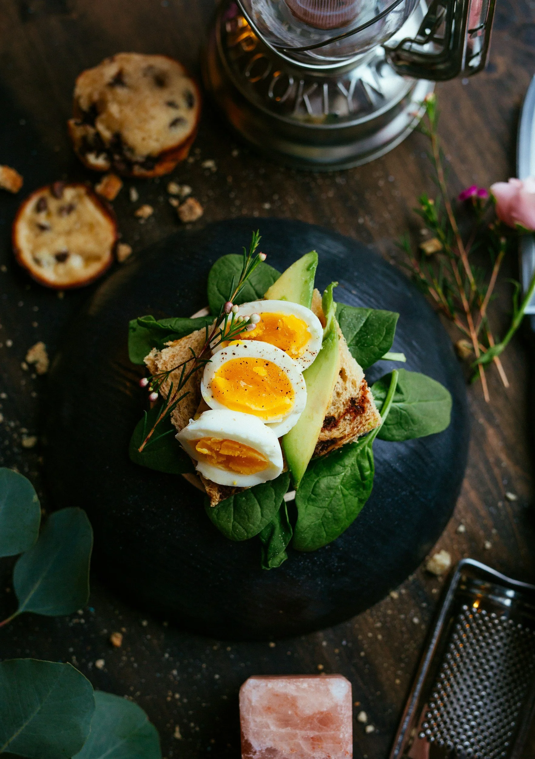 A plate with a breakfast toast topped with sliced soft-boiled eggs, avocado, and greens, on a dark surface with scattered cookie crumbs, a metal grater, a pink salt block, and some flowers nearby.