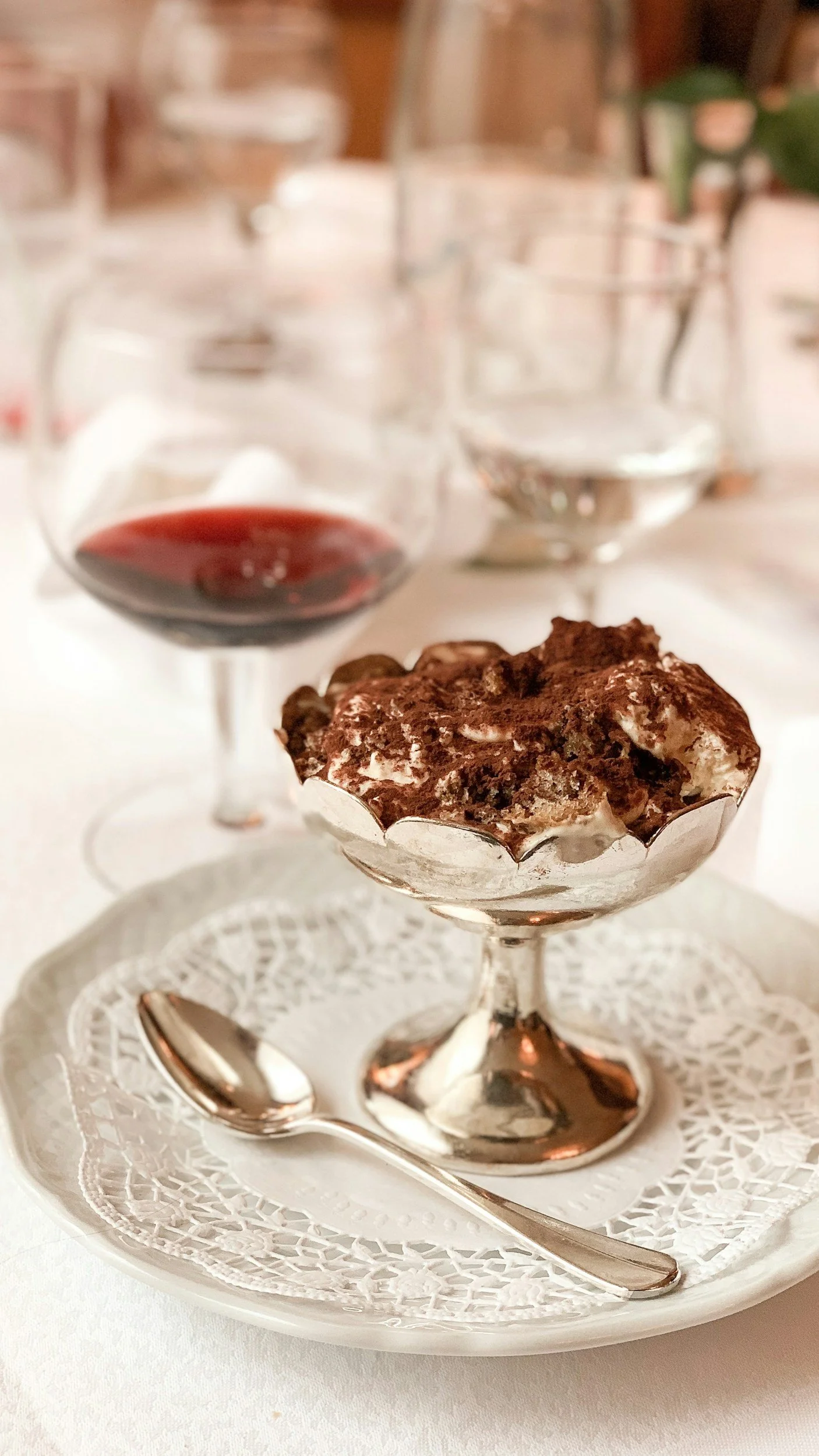 A dessert of tiramisu in an ornate glass dish on a white lace doily with a small spoon, and a glass of red wine and a glass of water in the background on a white table.