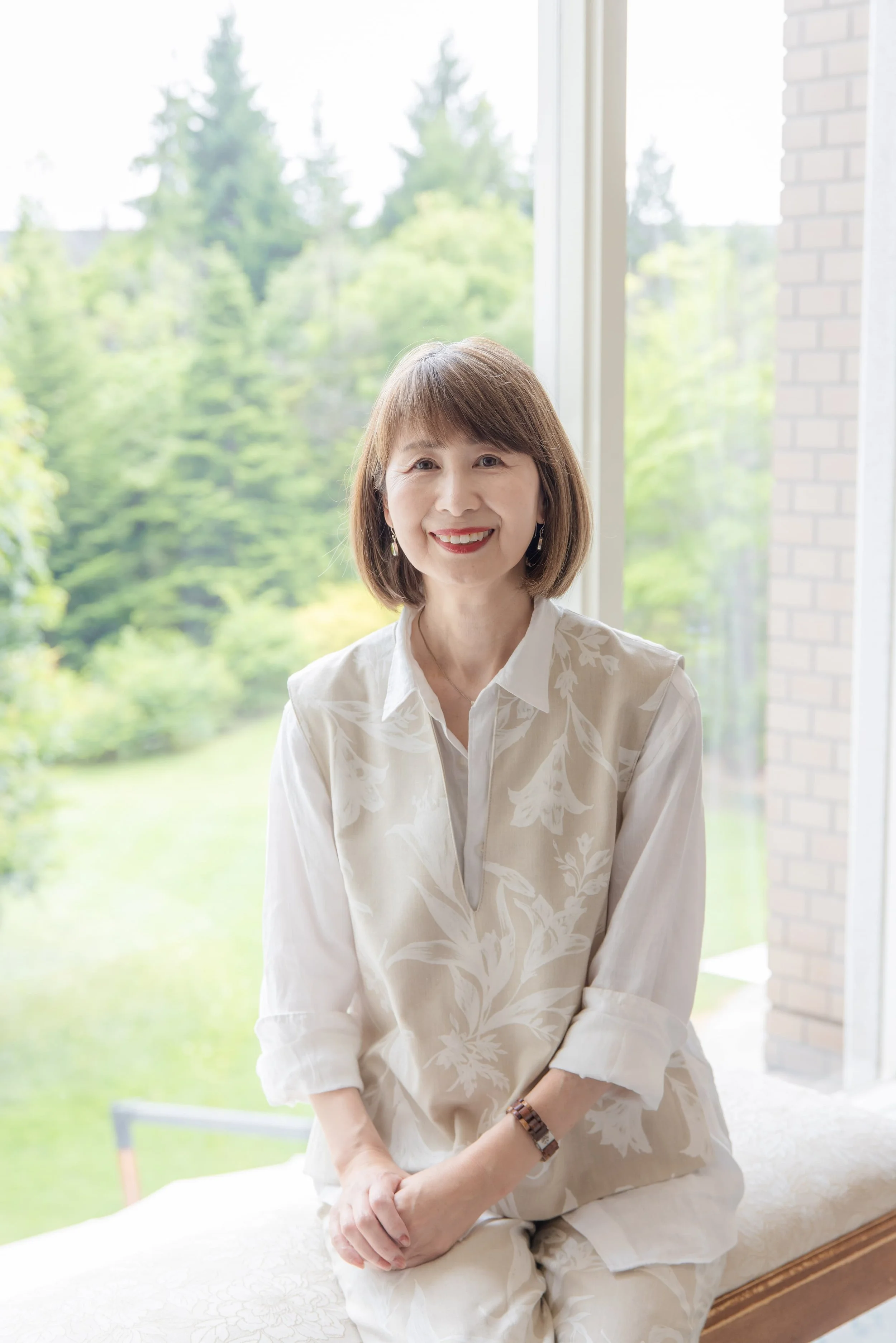 窓のそばに座る笑顔の中年女性。緑豊かな外の景色が背景。