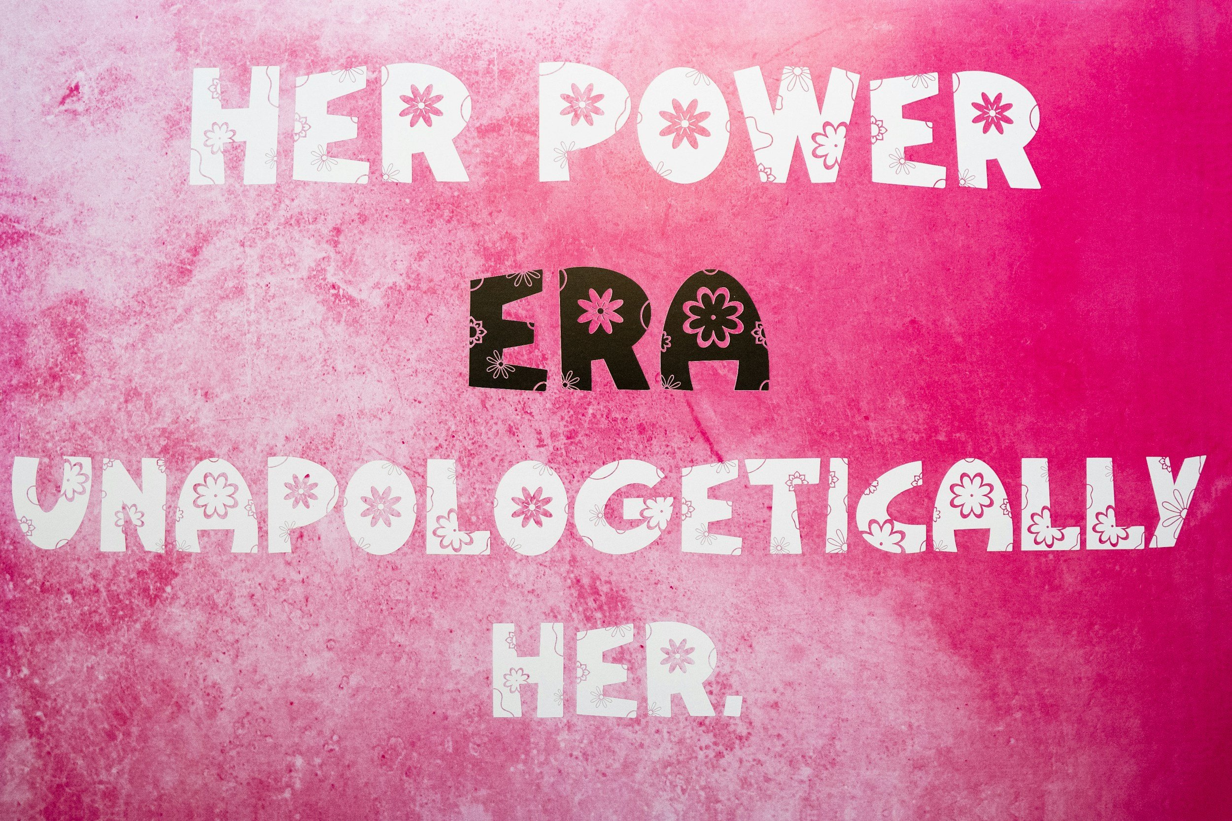 Pink background with the words 'Her Power Era' in white and black letters decorated with pink flowers.