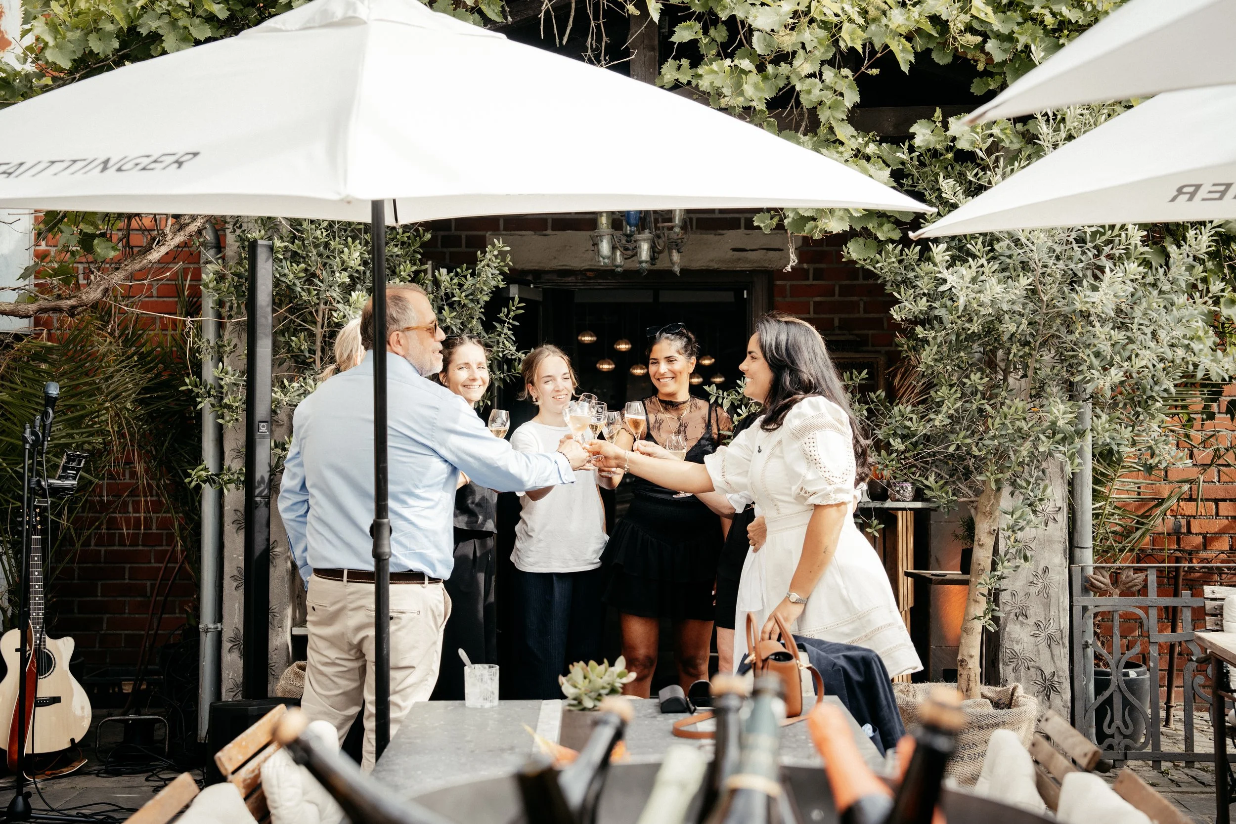Gruppe von sechs Menschen beim Anstoßen mit Gläsern beim Treffen im Außenbereich eines Restaurants oder Cafés, umgeben von Pflanzen und Sonnenschirmen.