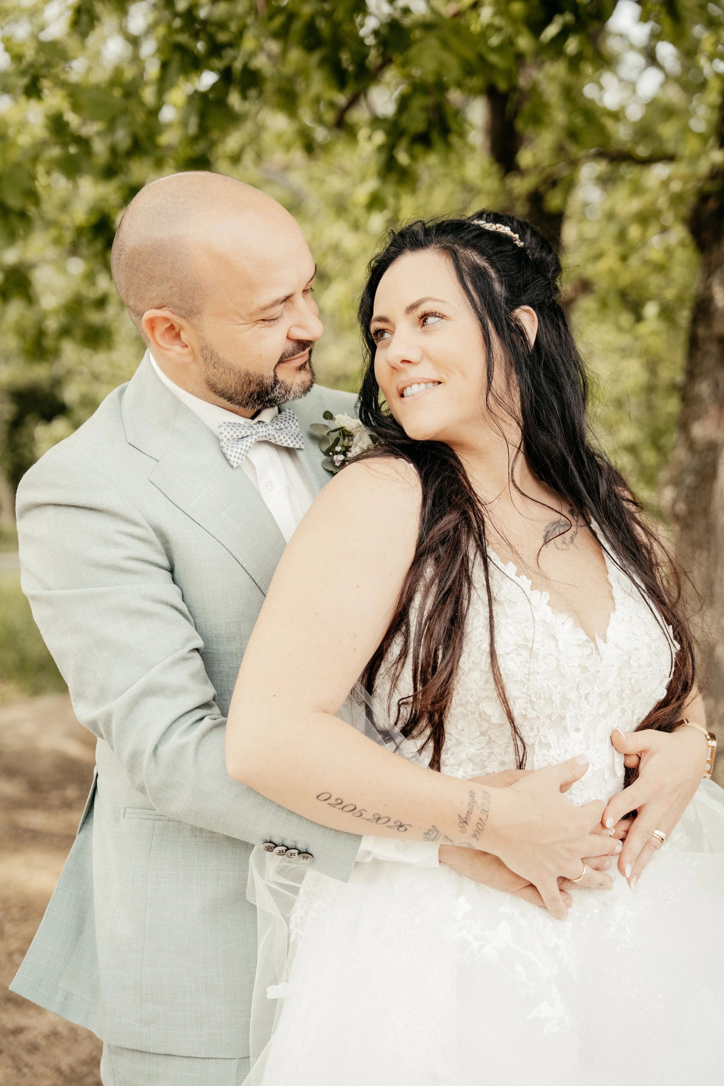 Ein Paar bei einer Hochzeit steht in einem Park, umarmt sich und lächelt sich liebevoll an.