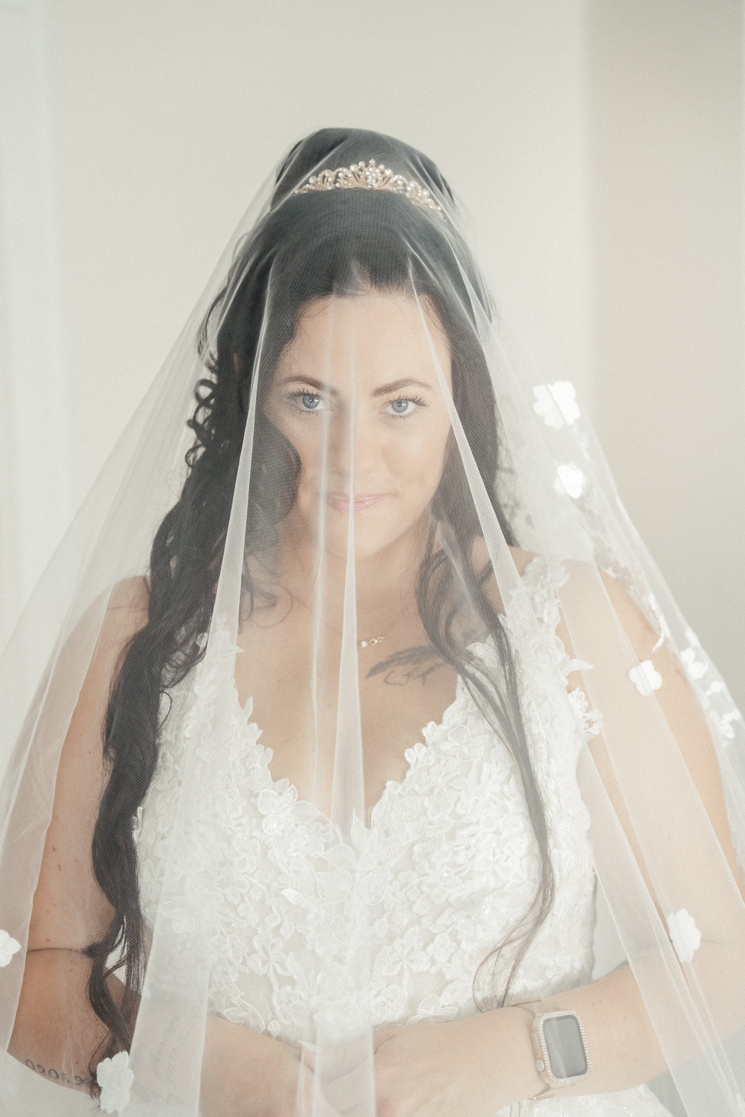 Braut in weißem Hochzeitskleid mit Spitze, mit Schleier und Diadem auf dem Kopf, lächelnd vor hellem Hintergrund.
