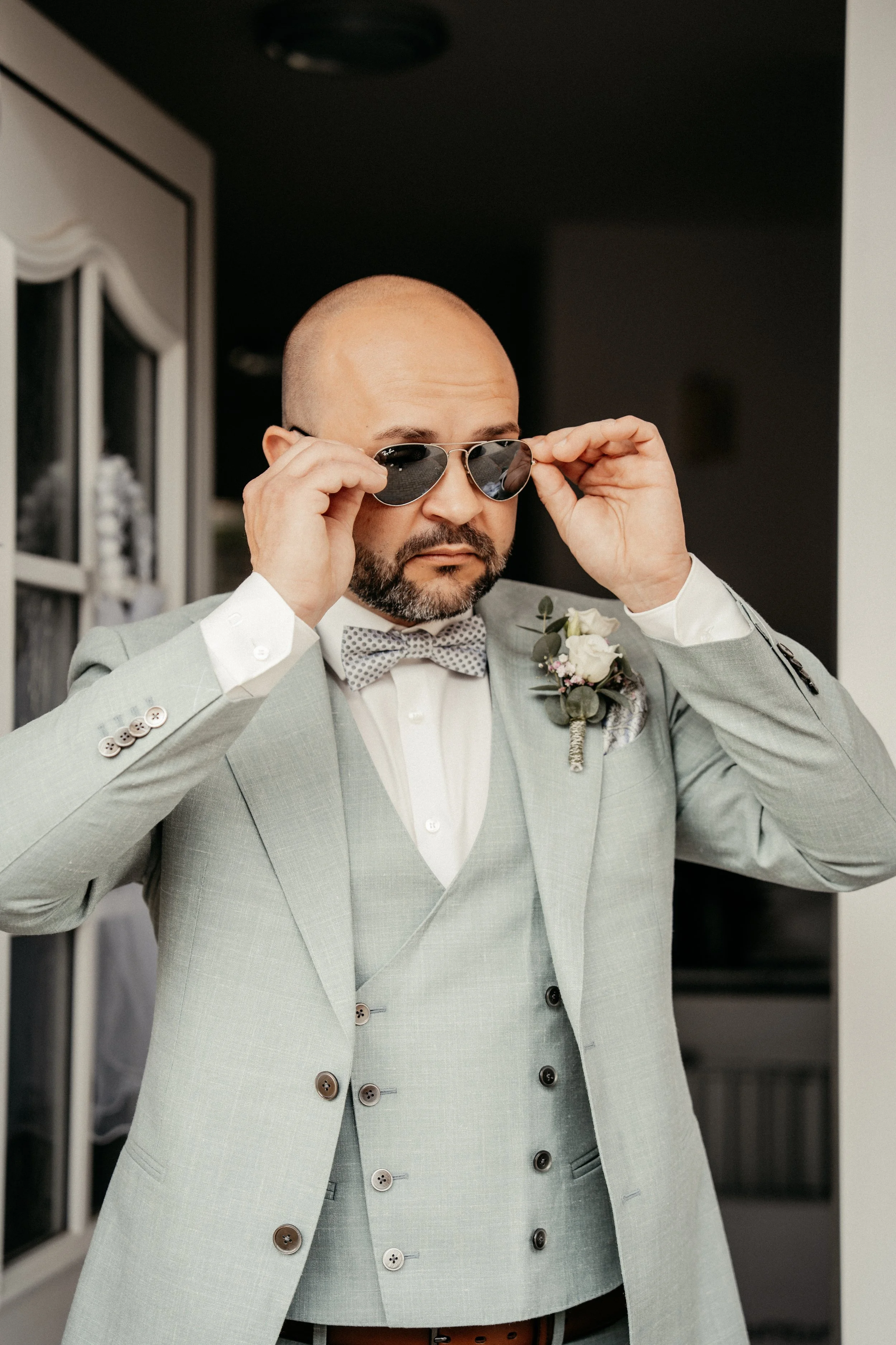 Ein Mann trägt eine helle Anzugjacke mit auffälligen Knöpfen, eine weiße Hemd mit Manschettenknöpfen, eine grau-weiße Fliege und eine Boutonniere, während er eine Sonnenbrille aufsetzt.