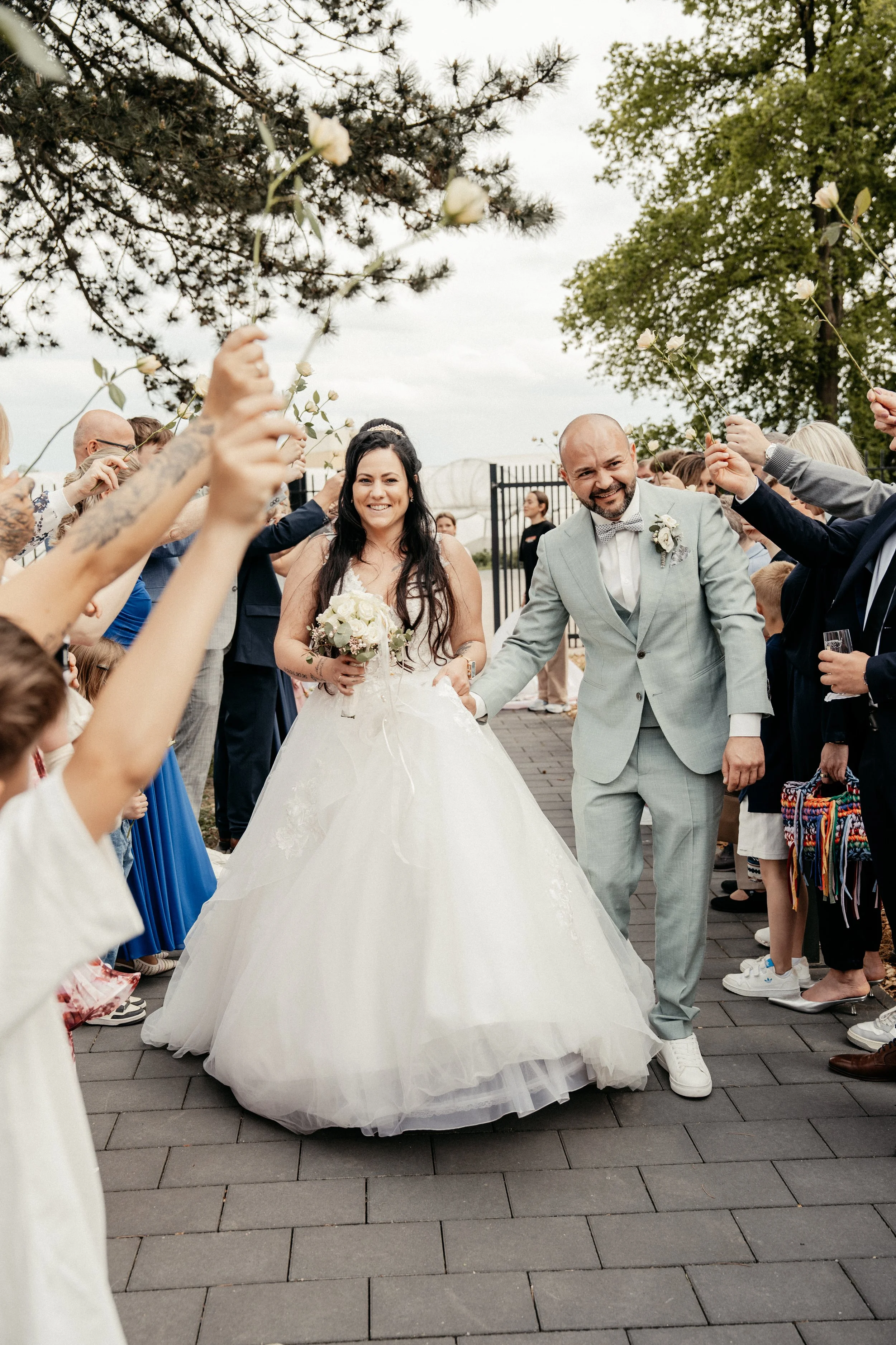 Braut und Bräutigam nach ihrer Hochzeit, umgeben von Gästen, beim Blumen-ritual, während sie sich die Hände reichen. Die Braut trägt ein weißes Brautkleid mit einem Tüllrock, der Bräutigam trägt einen hellgrauen Anzug mit Fliege. Gäste halten weiße R
