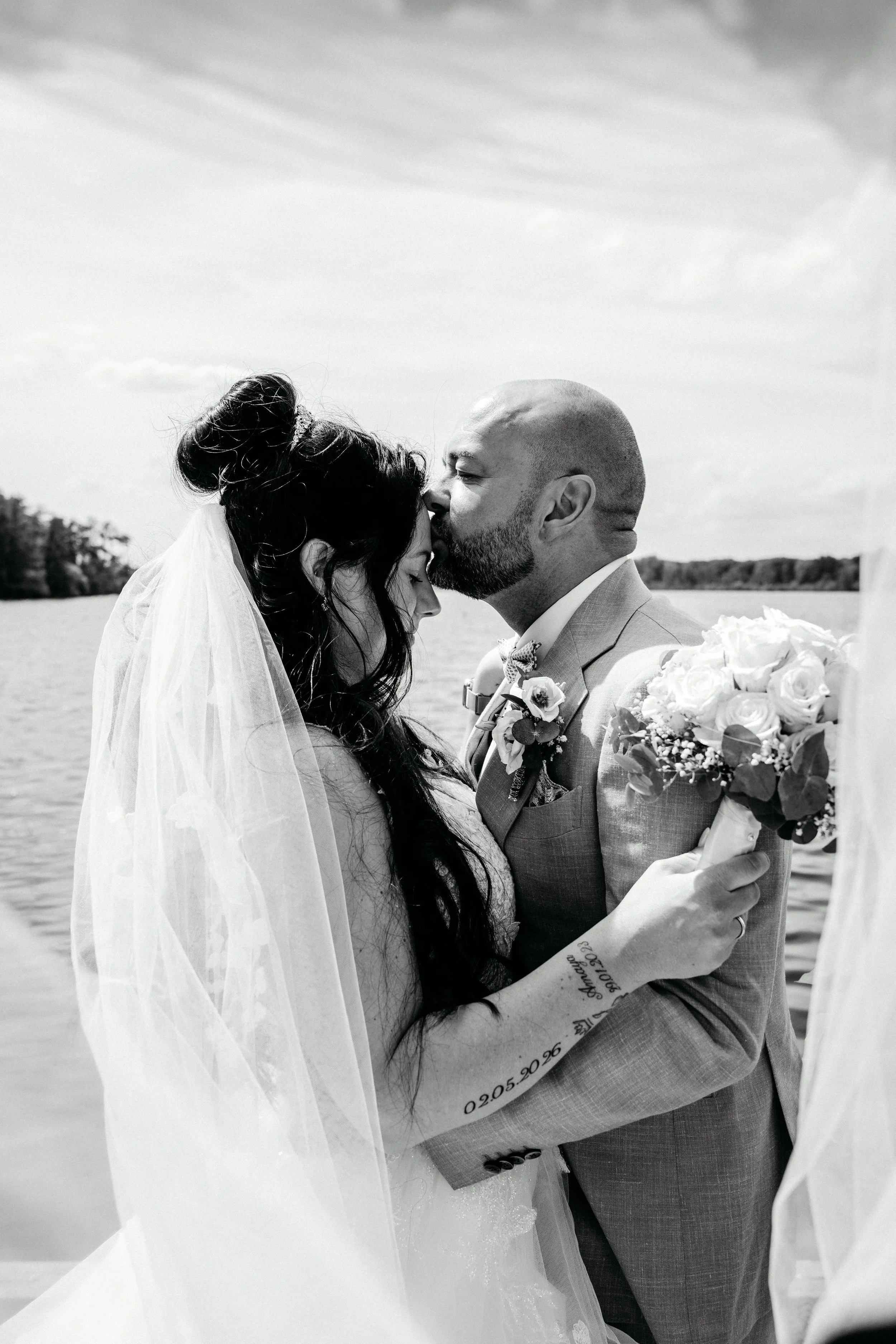 Braut und Bräutigam bei ihrer Hochzeit, die das Paar eng umarmt, mit Wasser im Hintergrund und Himmel über ihnen.